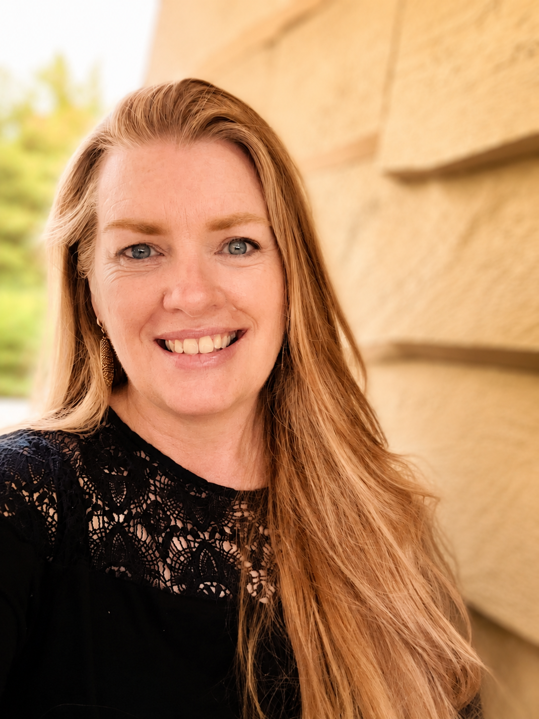 Close-up of smiling woman with long blonde hair, blue eyes, wearing a black lace top, earrings, standing outdoors near a stone wall with blurred greenery in the background.