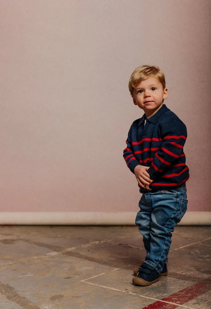 A young boy with blond hair, wearing a navy blue sweater with red and dark blue horizontal stripes, blue jeans, and black shoes, standing on a concrete floor against a neutral-colored wall.