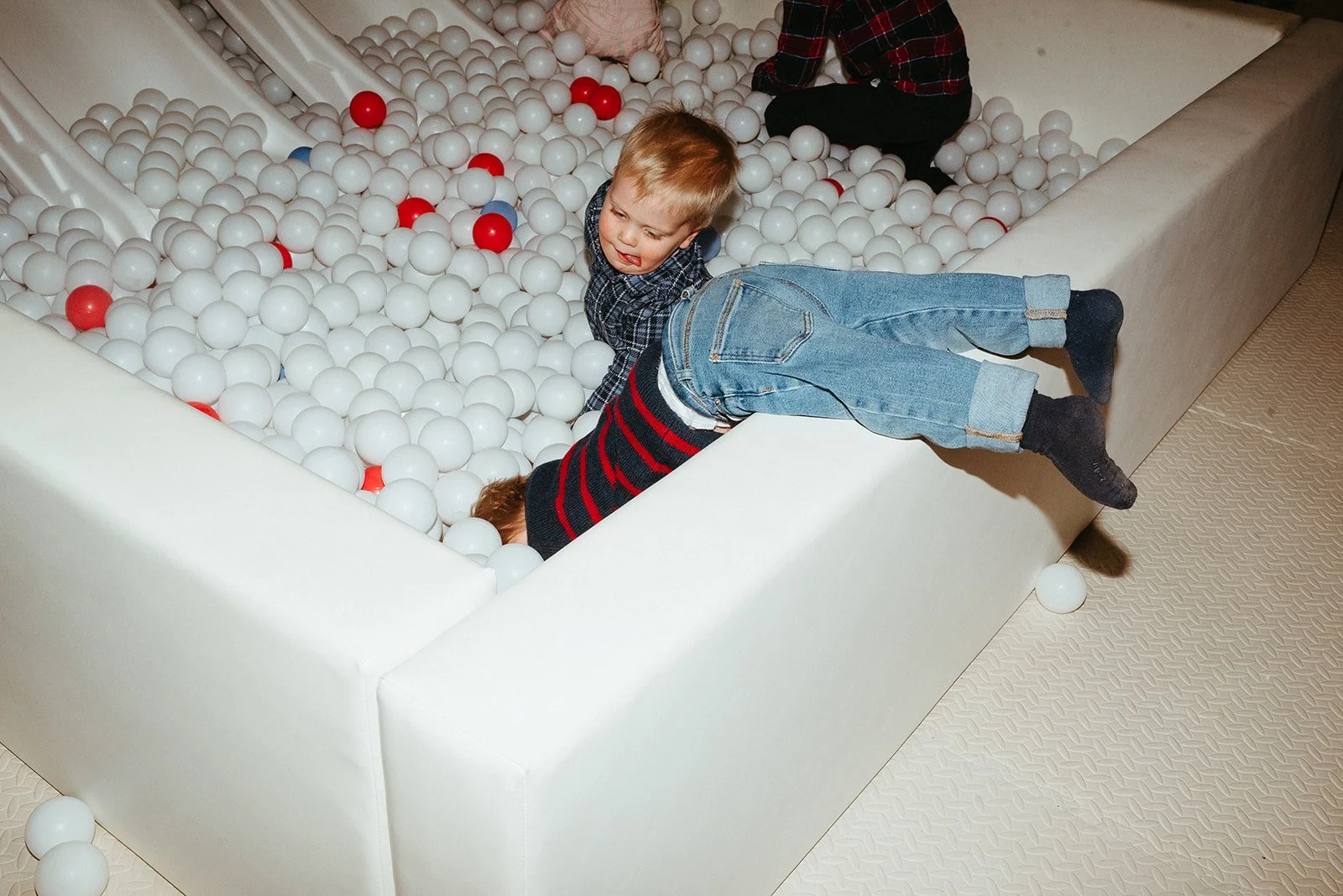 Two children playing in a ball pit filled with white and red plastic balls, with one child hanging over the edge and the other climbing in.
