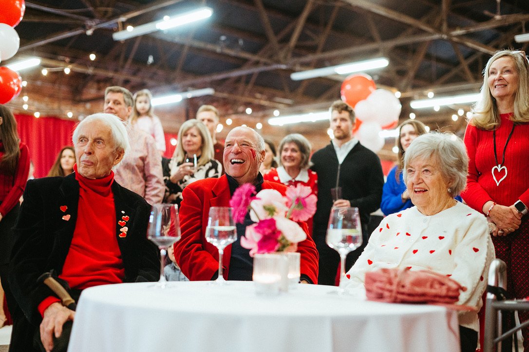 Elderly man and woman sitting at a table with glasses and a flower arrangement, surrounded by smiling people at a celebration with red, white, and pink decorations.