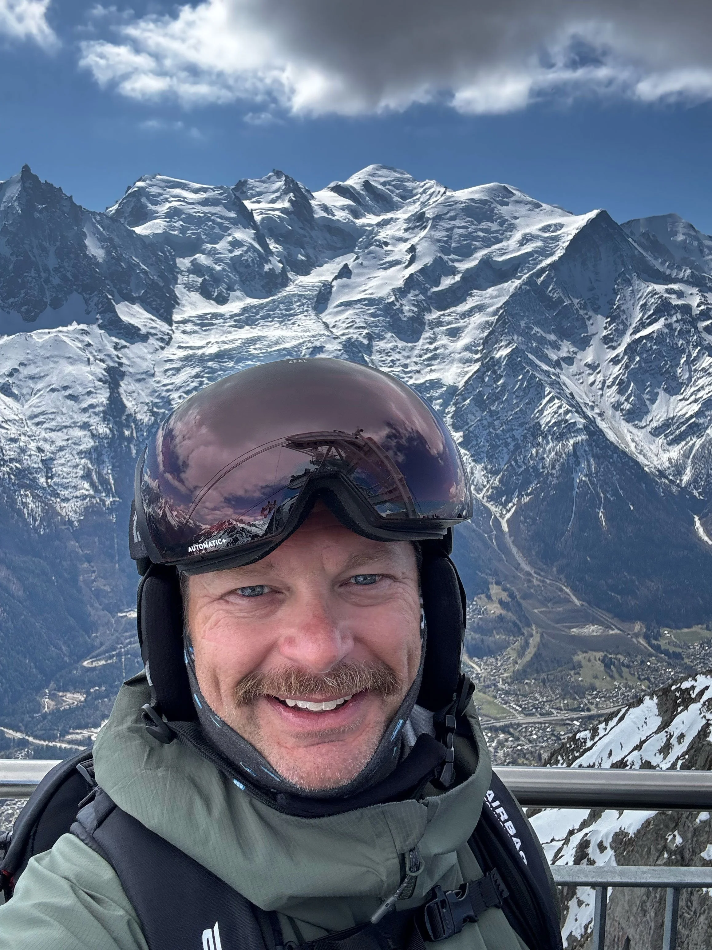 A man wearing a helmet and goggles smiling in front of snow-capped mountains during a ski or snowboarding trip.