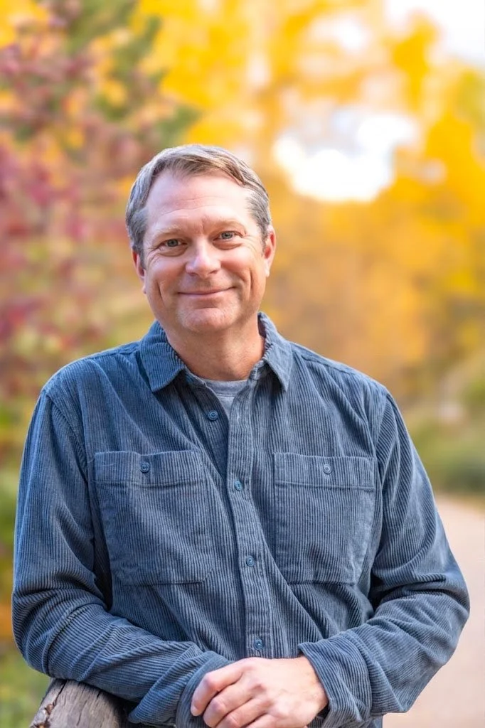 Dan Sherman standing outdoors in Aspen, Colorado in the fall, with blurred autumn trees with yellow and orange leaves in the background.