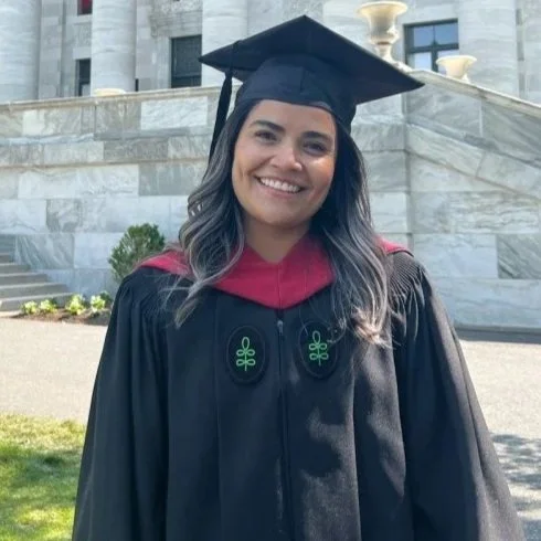 Woman in graduation cap and gown standing outdoors in front of a building with stairs and plants, smiling.