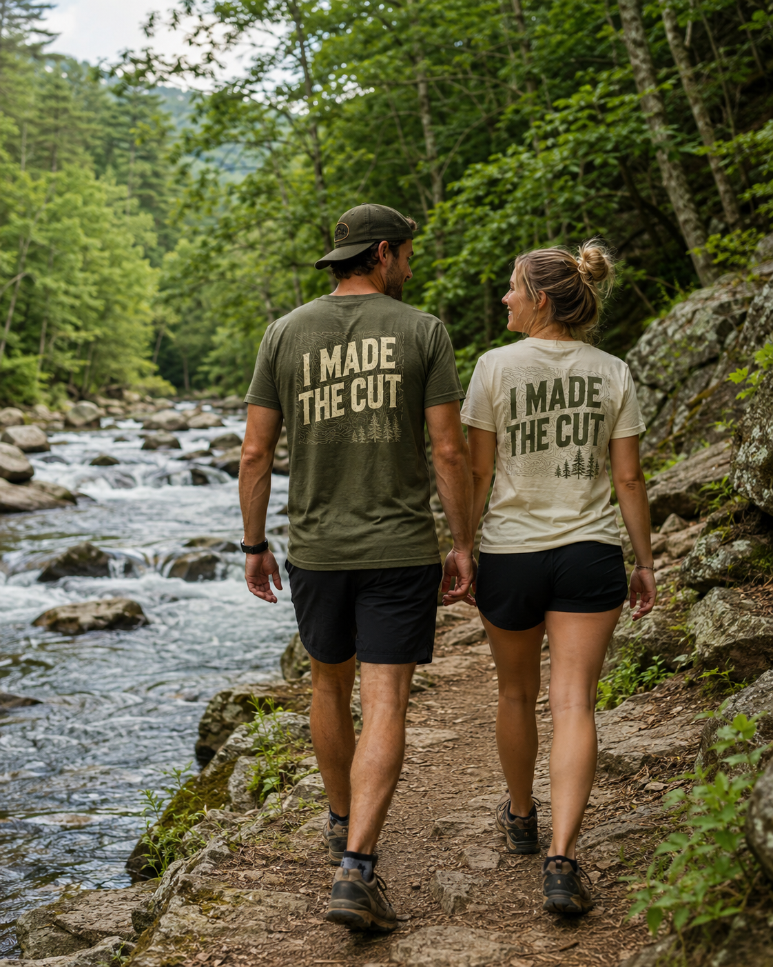 A couple walking hand in hand on a trail beside a creek in a forest, wearing matching shirts that say 'I MADE THE CUT' with trees and topographical lines on them.