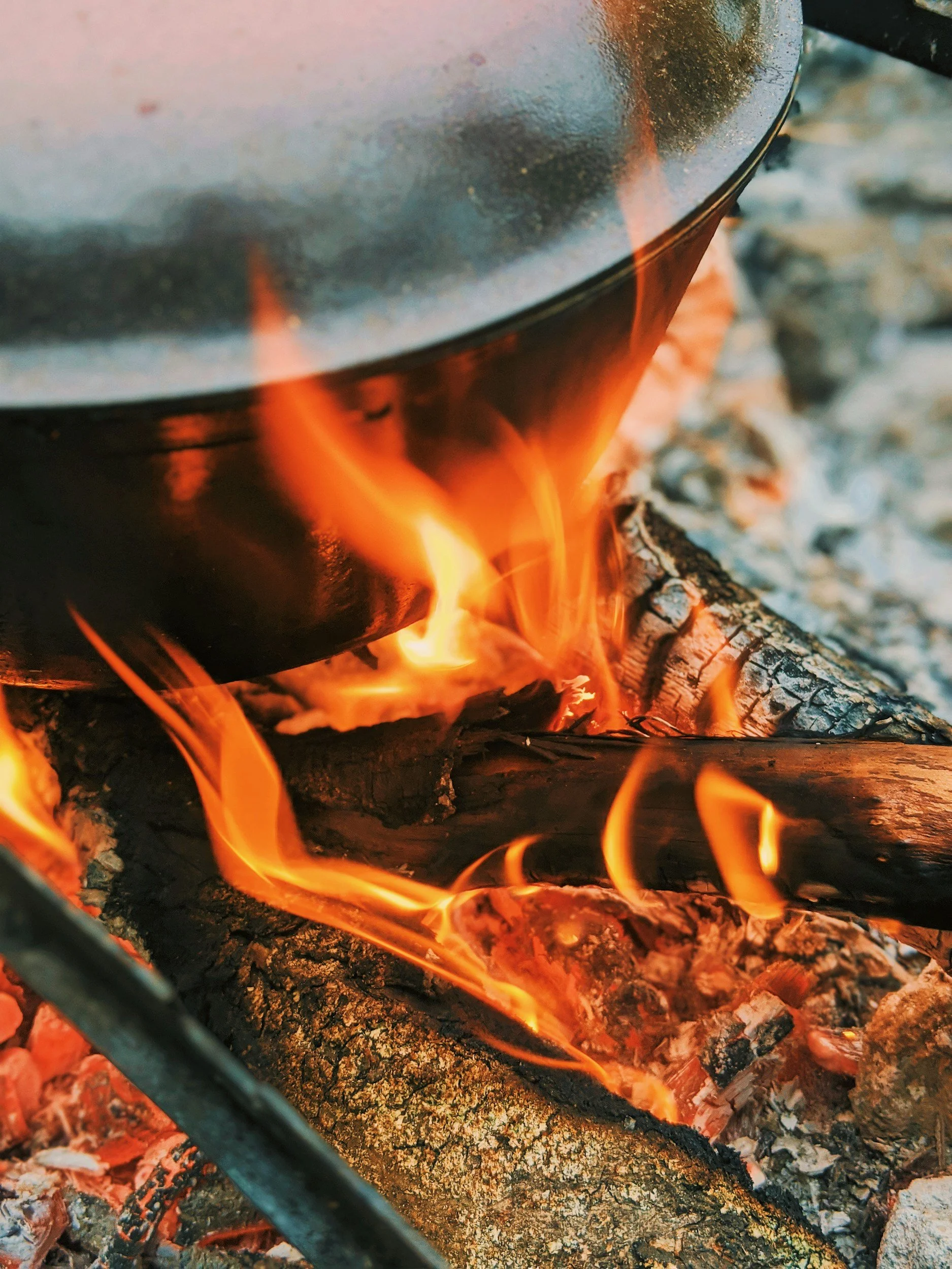 Close-up of a fire in a campfire with glowing orange flames and burning logs, with ashes and small embers visible at the base.