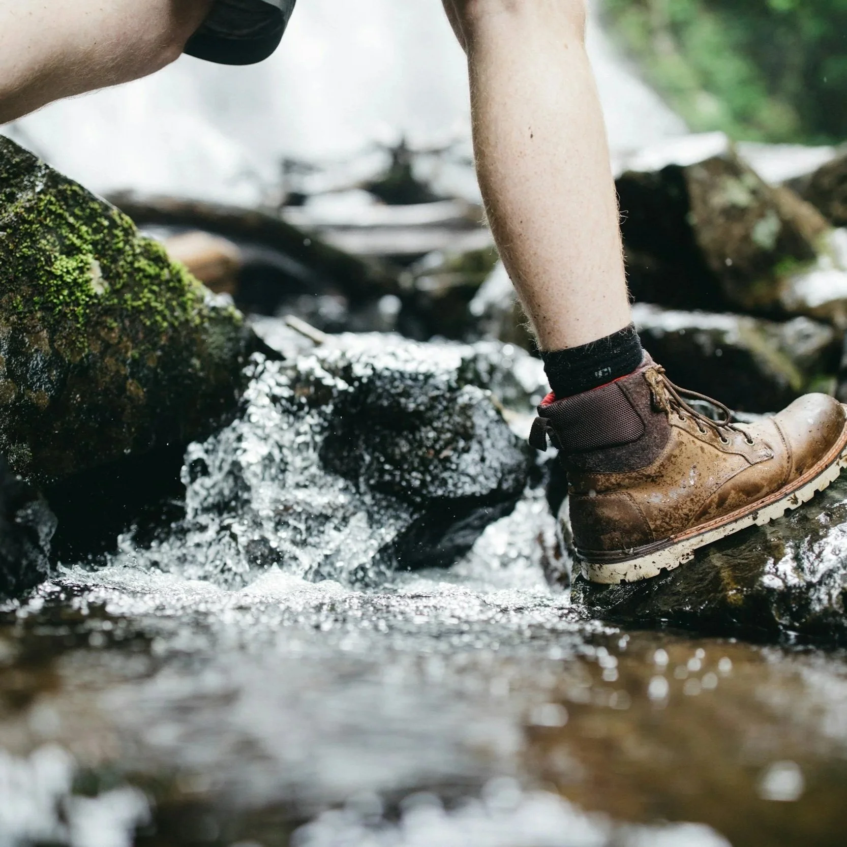 Close-up of a person crossing a stream on rocky terrain, wearing hiking boots and camouflage shorts, with water flowing around their feet.