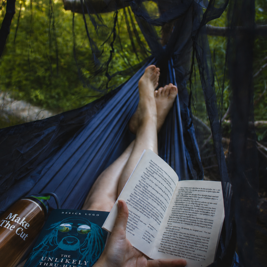 Person relaxing in a hammock with a book and a water bottle in a green outdoor setting.