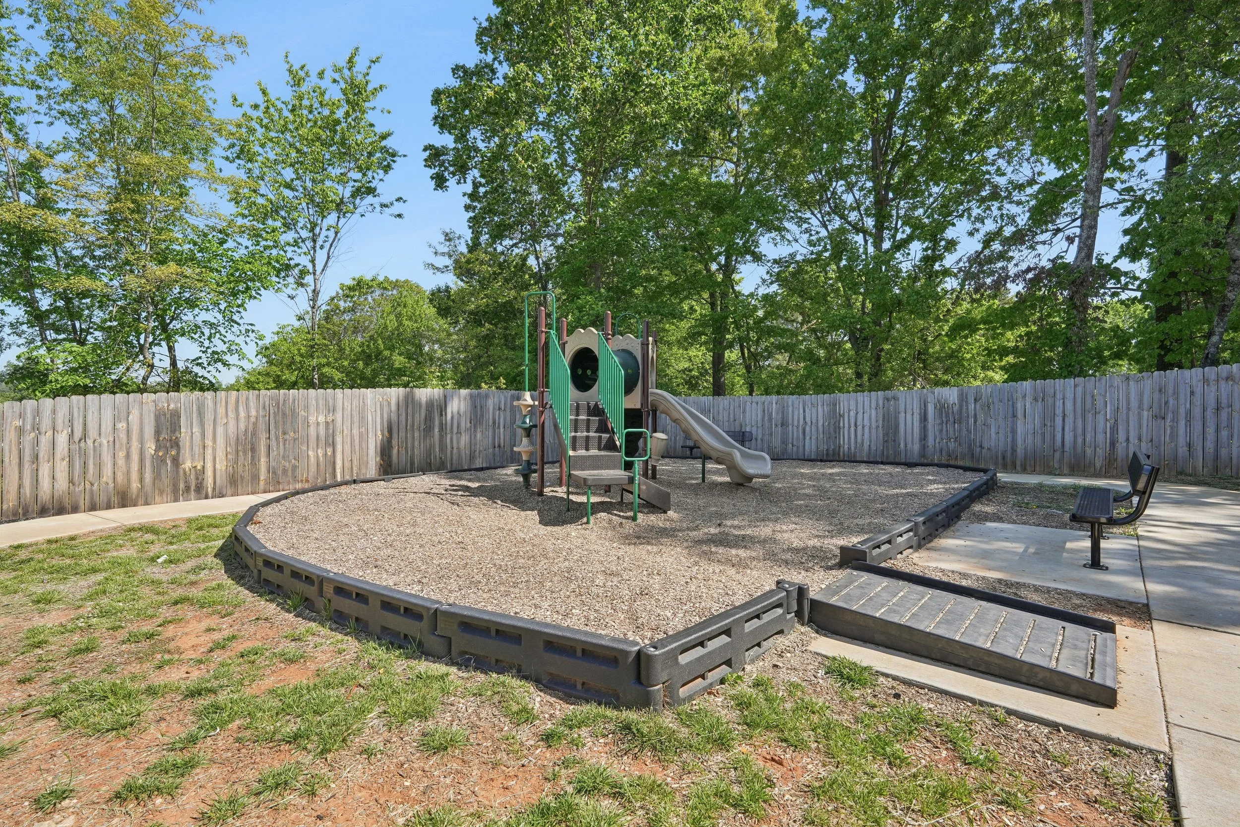 A small playground with a slide and climbing structure in a fenced area, surrounded by trees and benches on a sunny day.