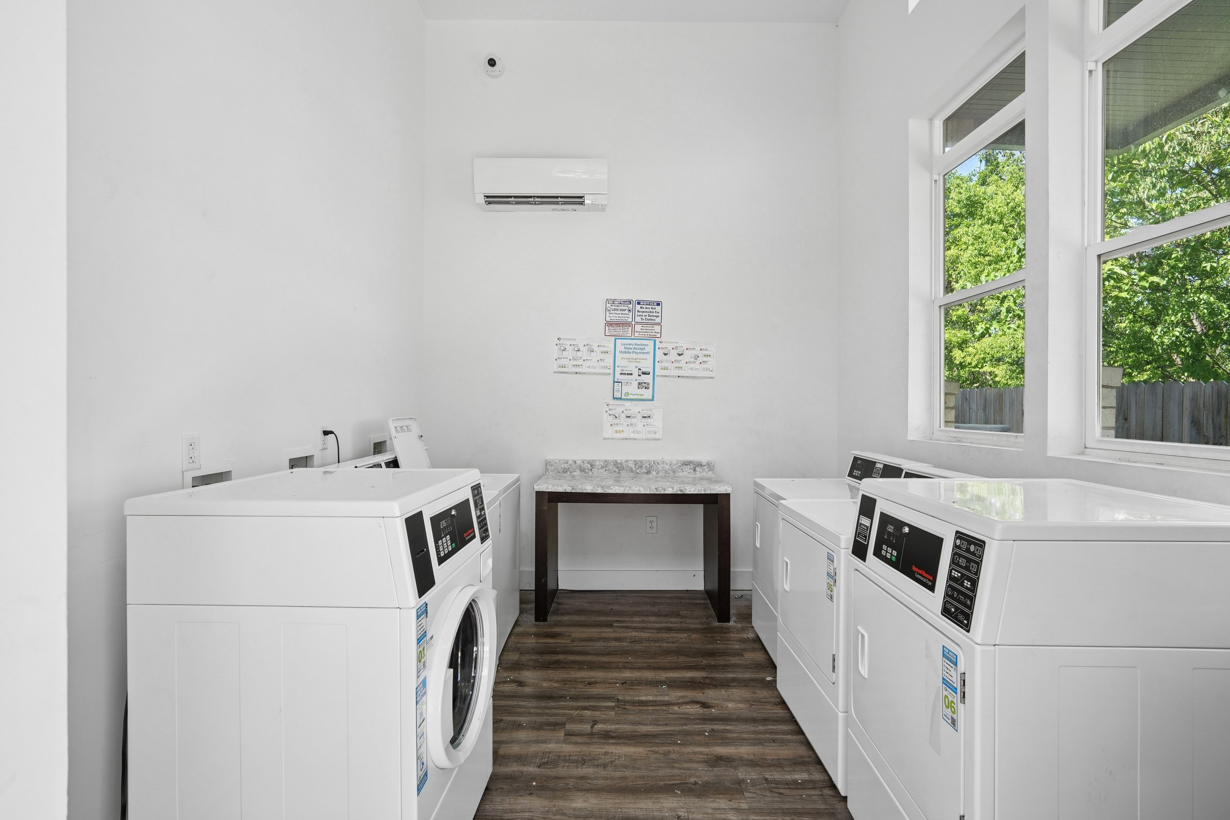 Laundry room with four white washing machines, a granite-top table in the center, a large window letting in natural light, and instructional signs on the wall.