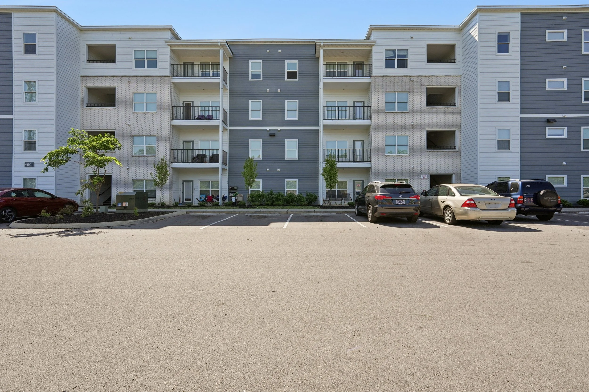 Exterior view of a modern, multi-story apartment building with white and gray siding, small balconies, and parking lot with cars in front.