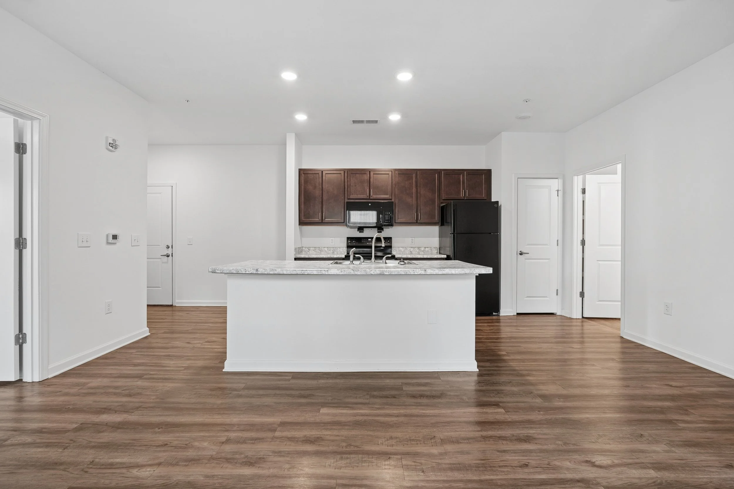 Empty modern kitchen with brown cabinets, black appliances, granite countertops, white walls, and wood flooring.