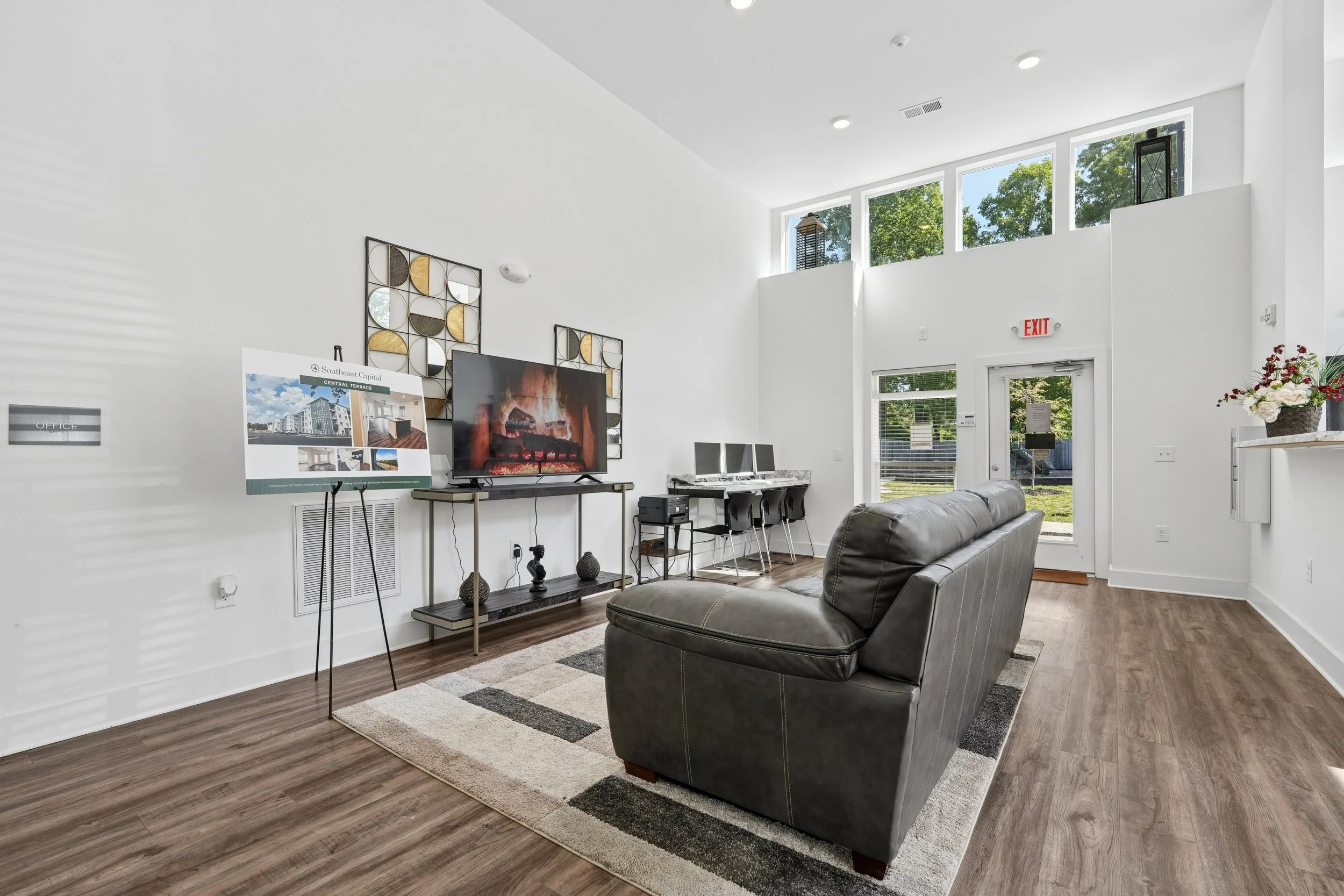 A modern lobby area with white walls, large windows, a leather couch, a TV, a display board, and decorative wall art.