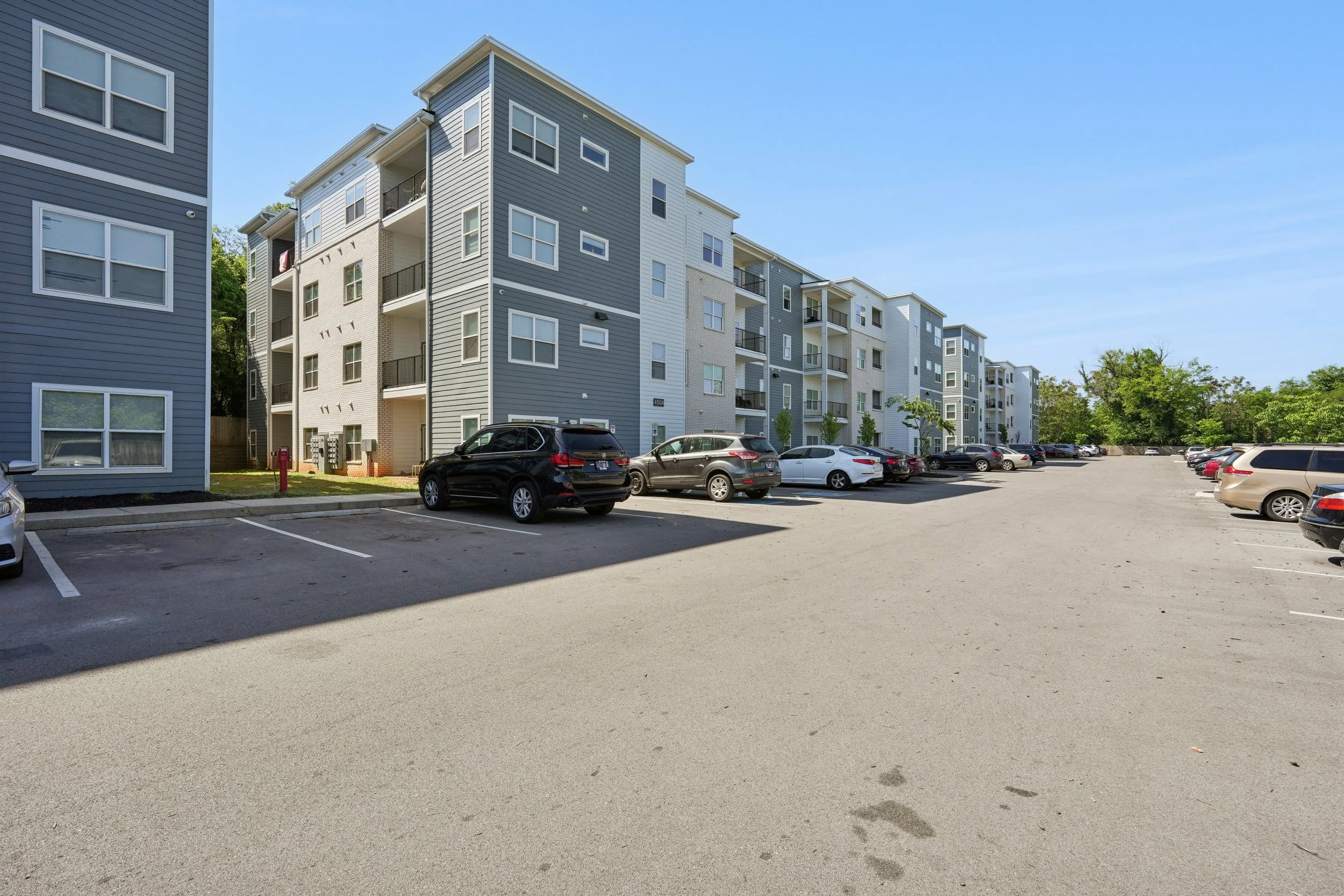 Multistory apartment complex with gray and white exterior, balconies, and numerous windows. Parking lot with several cars in front of the building, and a clear blue sky overhead.