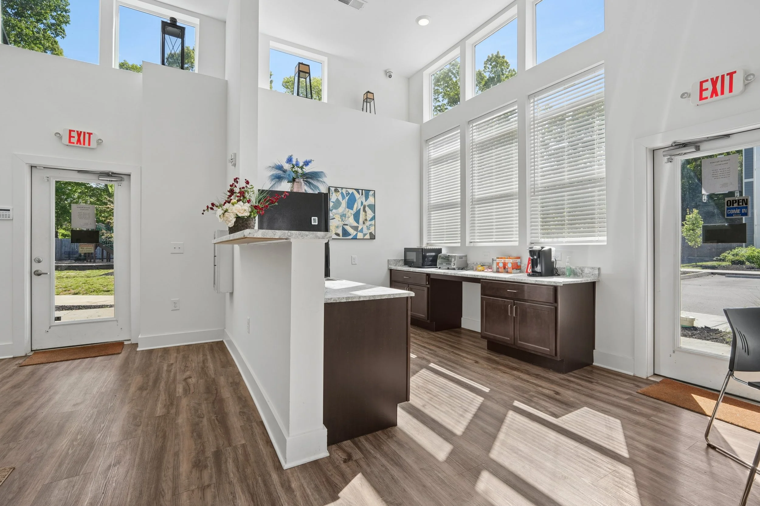 Interior of a modern commercial kitchen with white walls and large windows, brown cabinets, a marble countertop, and a coffee machine on the counter.