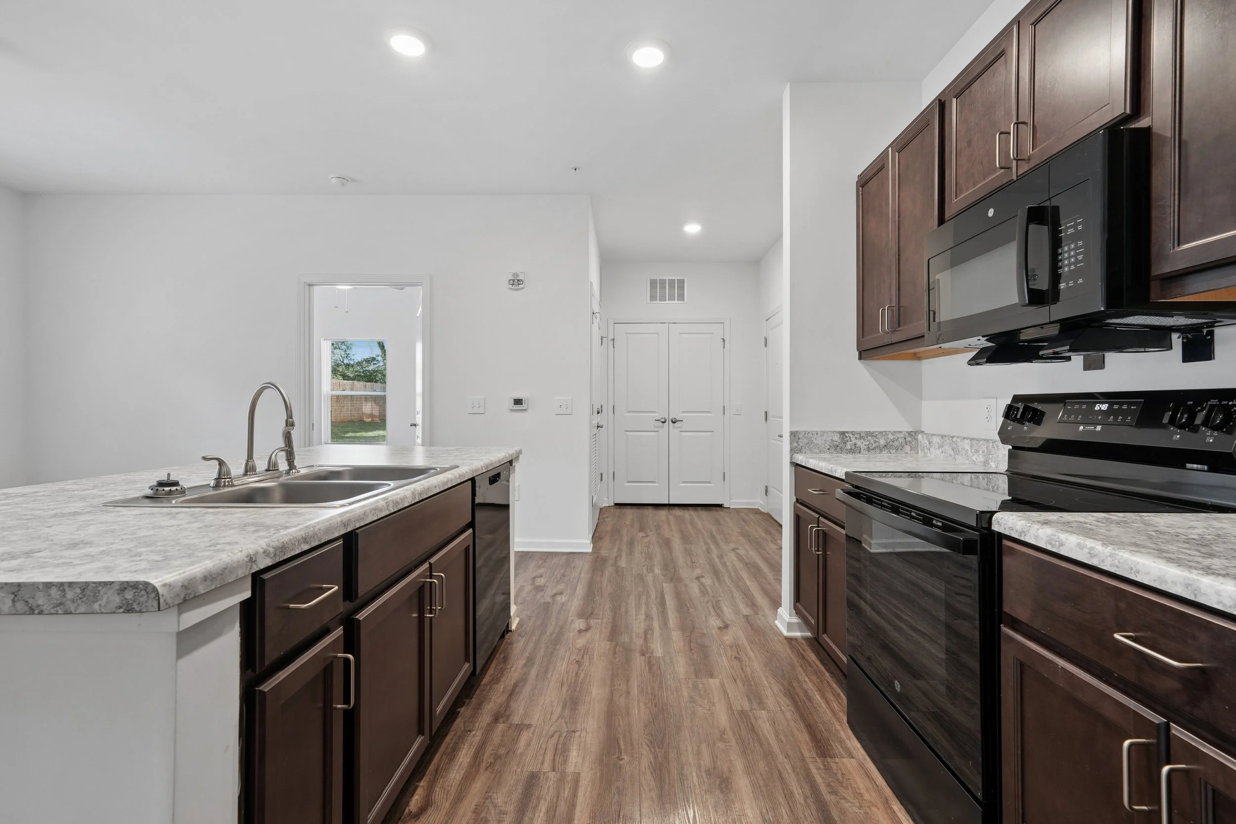 Kitchen with dark brown cabinets, marble countertops, stainless steel sink, black microwave, electric stove, white walls, and wooden flooring.