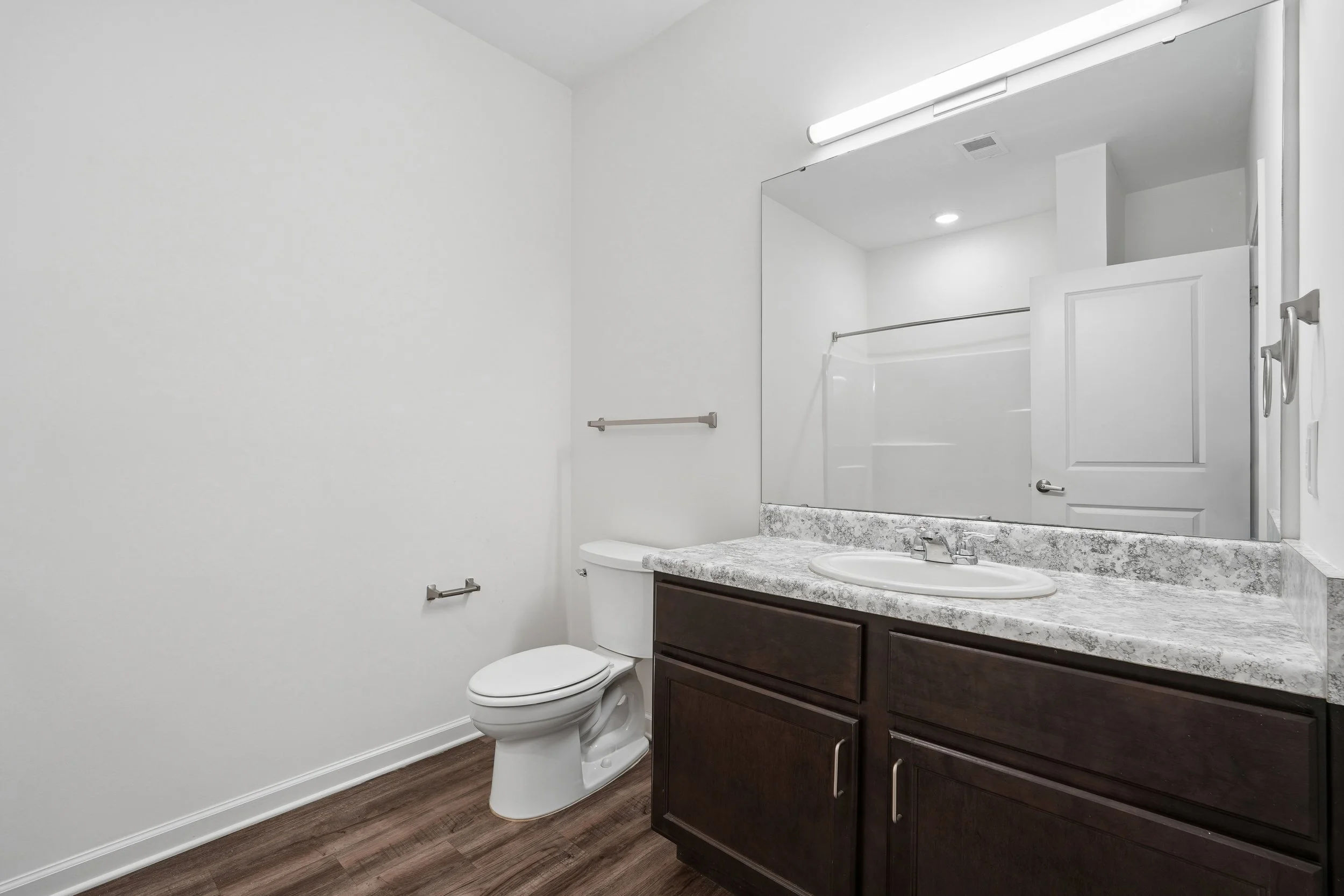 A modern bathroom with white walls, a toilet, dark wooden vanity with a granite countertop, a sink, a large mirror, a picture light above the mirror, a towel bar next to the toilet, and a ceiling vent.