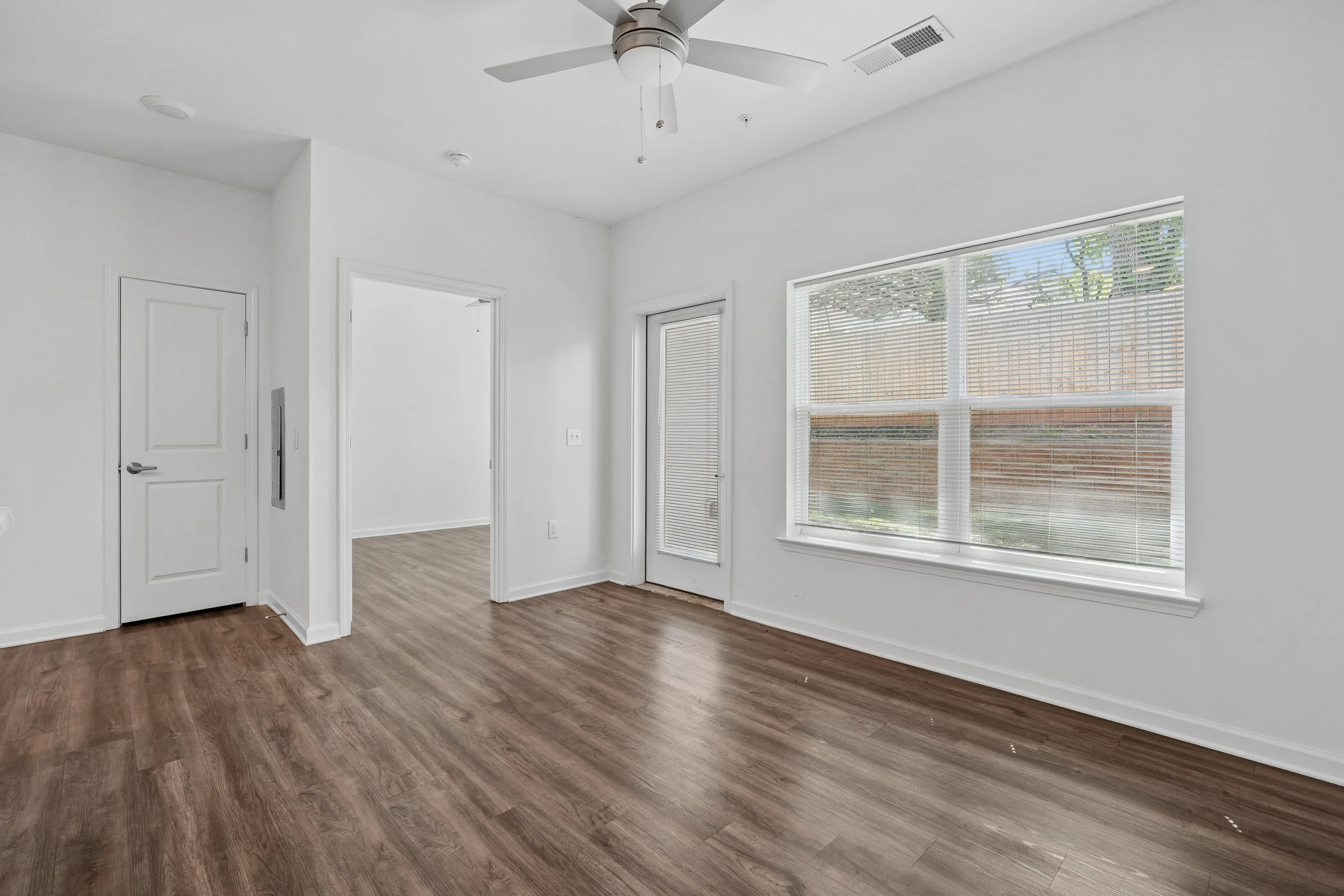 Empty room with white walls, large front and side windows with blinds, light brown wood flooring, ceiling fan, and a door.