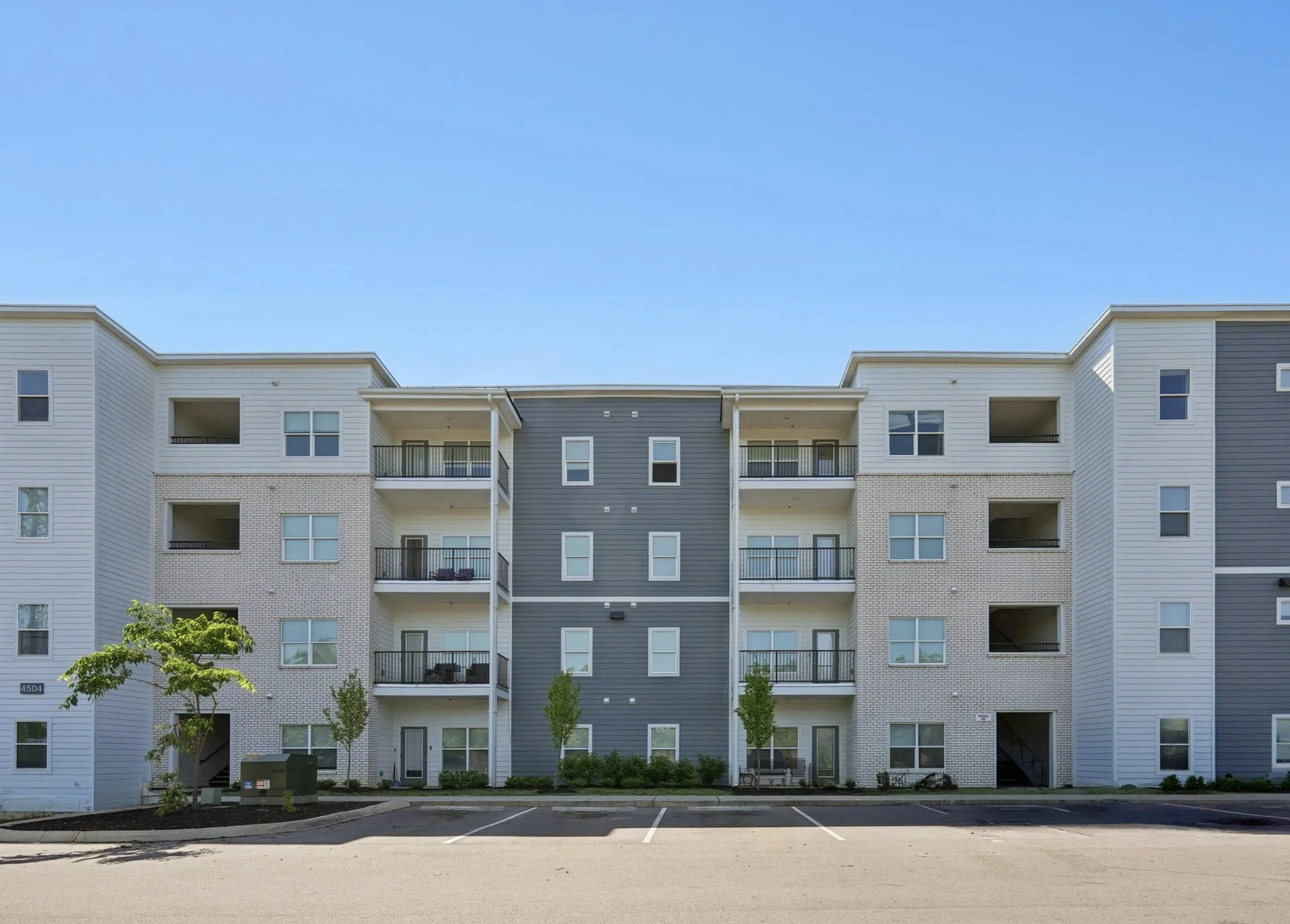 A modern multi-story apartment building with white and gray exterior walls, balconies, and windows, with small trees and an empty parking lot in front, under a clear blue sky.