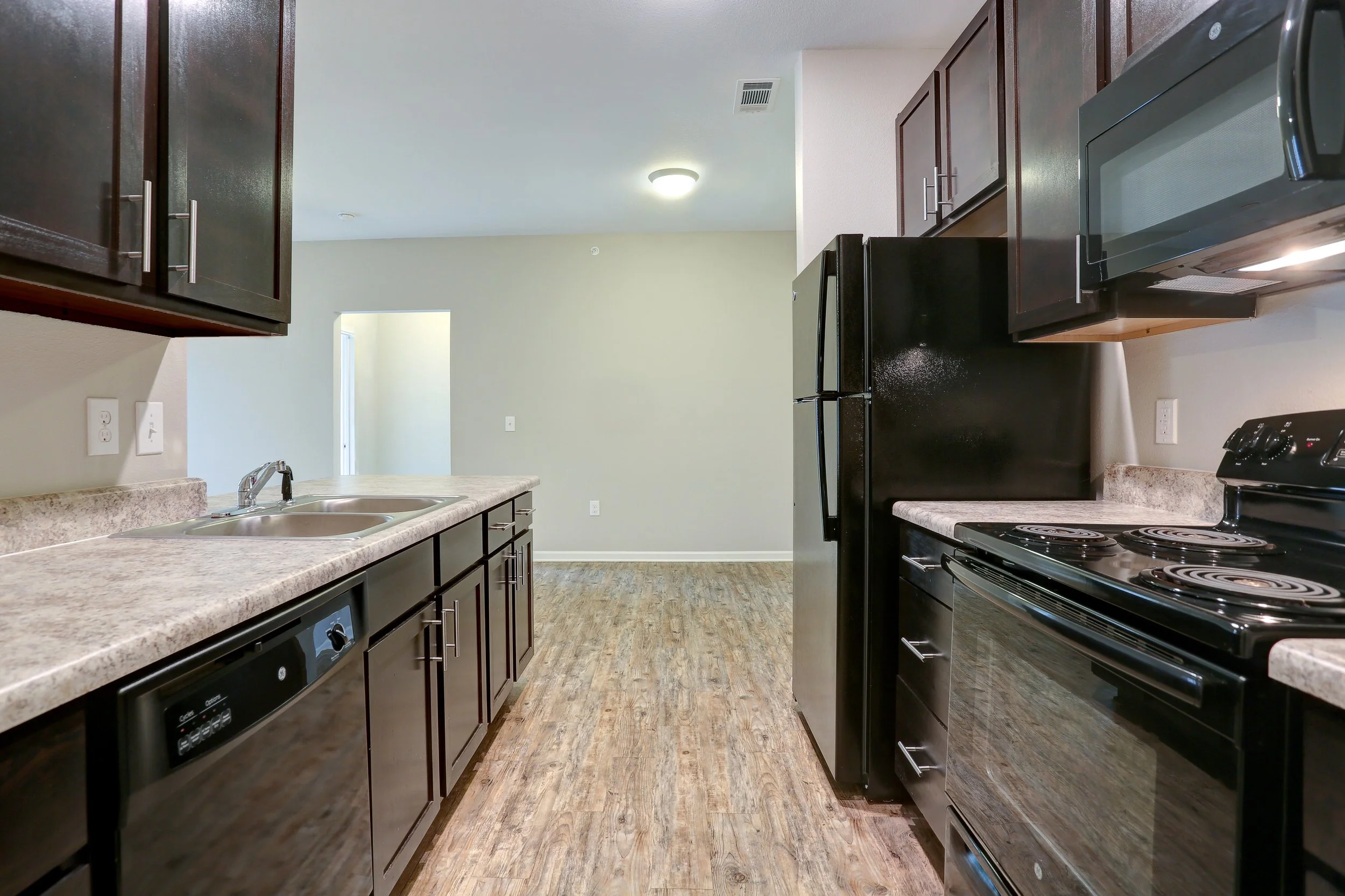 Kitchen with dark wood cabinets, beige countertops, black appliances including a stove, refrigerator, and microwave, stainless steel sink, and wood-look flooring, with light-colored walls and a ceiling light.