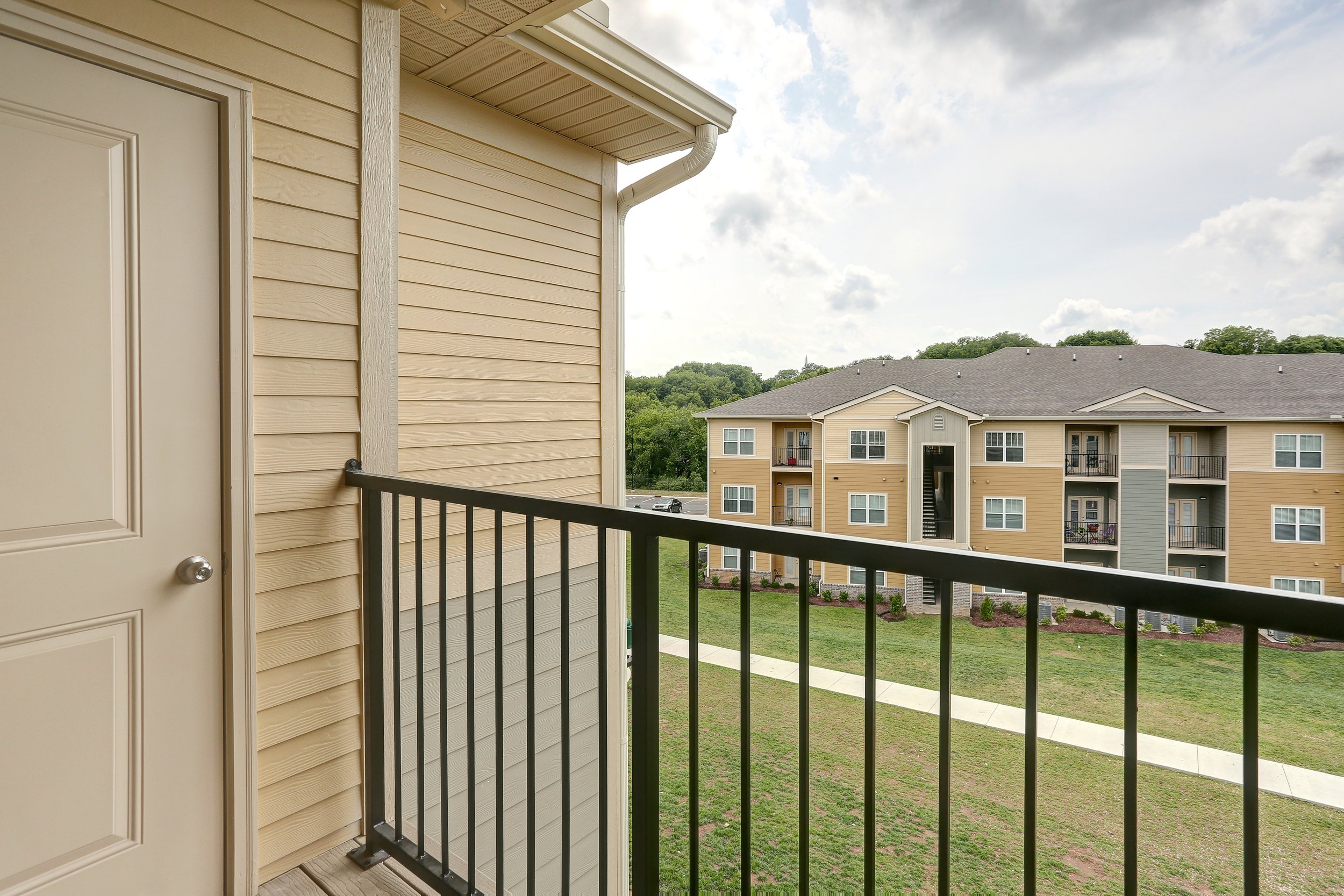 View from a balcony showing neighboring apartment buildings with beige and grey siding, multiple windows, and balconies, under a partly cloudy sky.