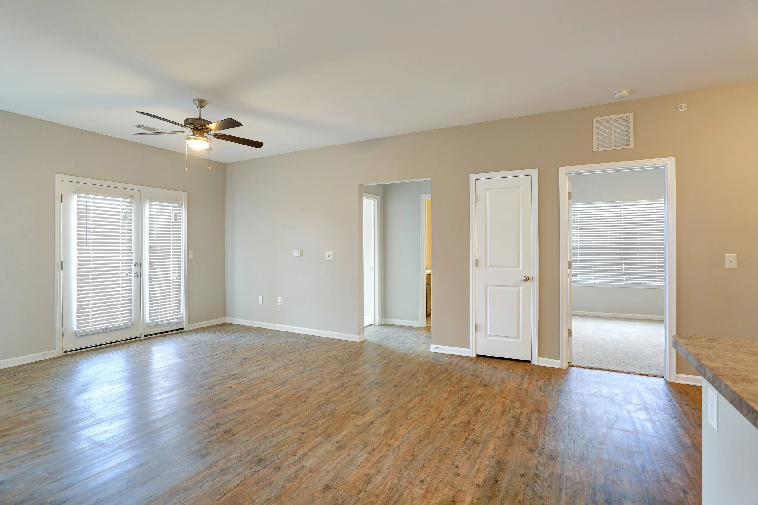 Interior of a spacious living area with white walls, wooden flooring, and multiple doors and windows with blinds. A ceiling fan with a light is mounted on the ceiling.