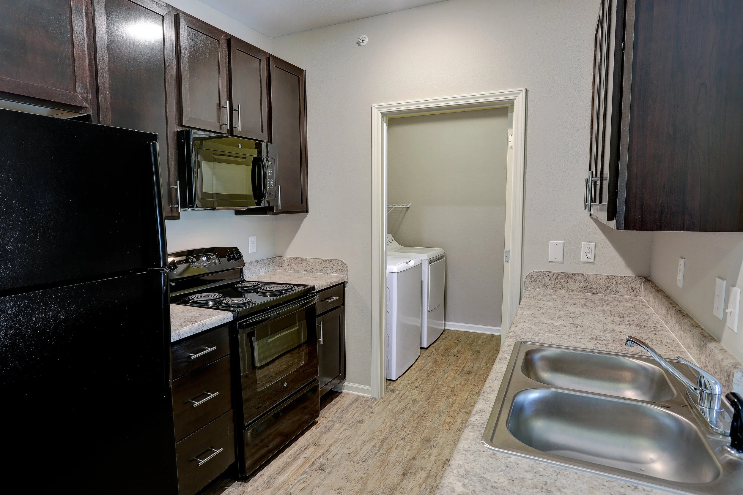 Kitchen with dark brown cabinets, black refrigerator, stove, microwave, beige countertops, stainless steel sink, and laundry area with washing machine in a small doorway.
