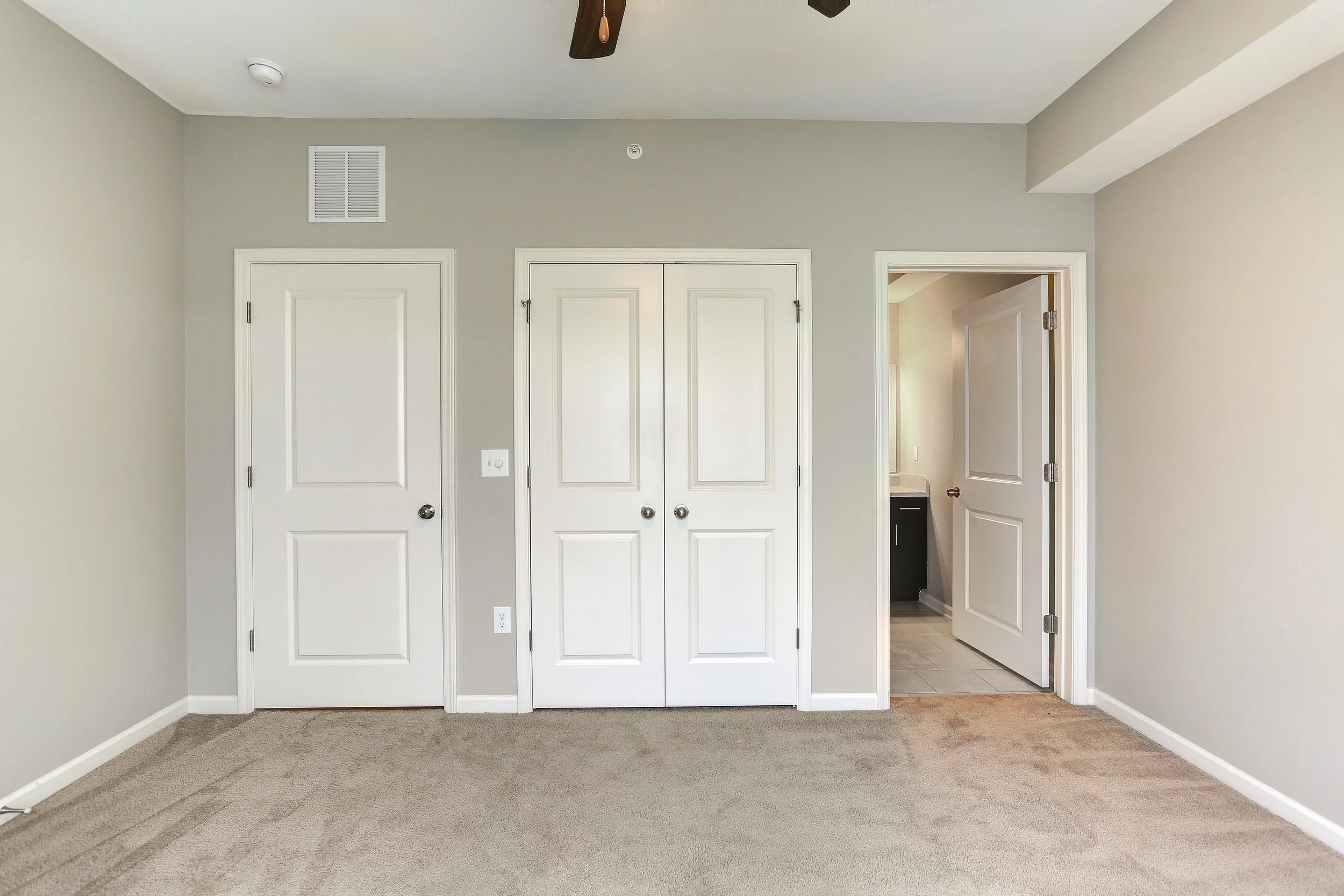Empty bedroom with beige carpet, light gray walls, closed closet doors, and an open door showing a bathroom or another room.