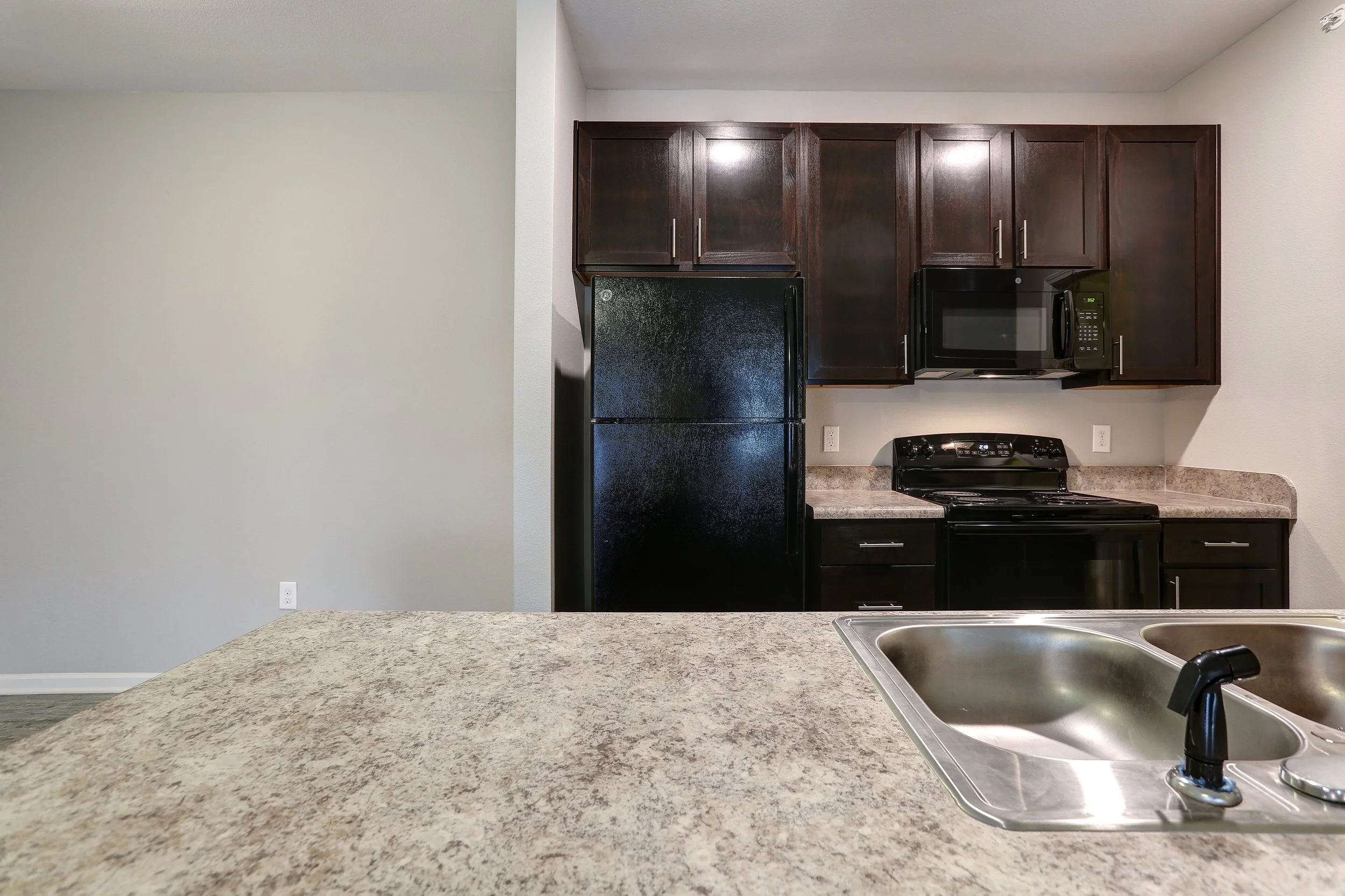 Kitchen with granite countertop, black refrigerator, microwave, oven, dark wooden cabinets, and a double sink.