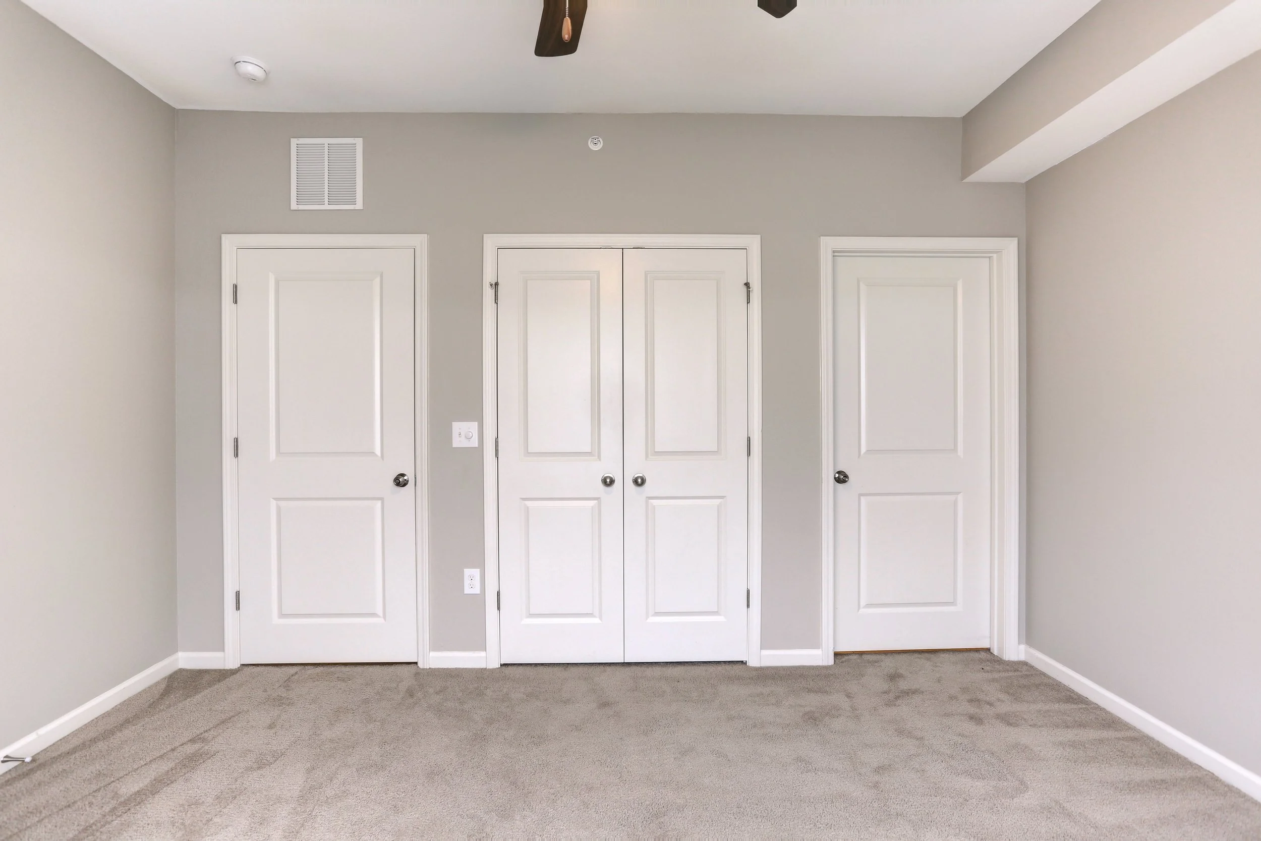 Interior view of beige carpeted room with three closed white doors and a ceiling fan.