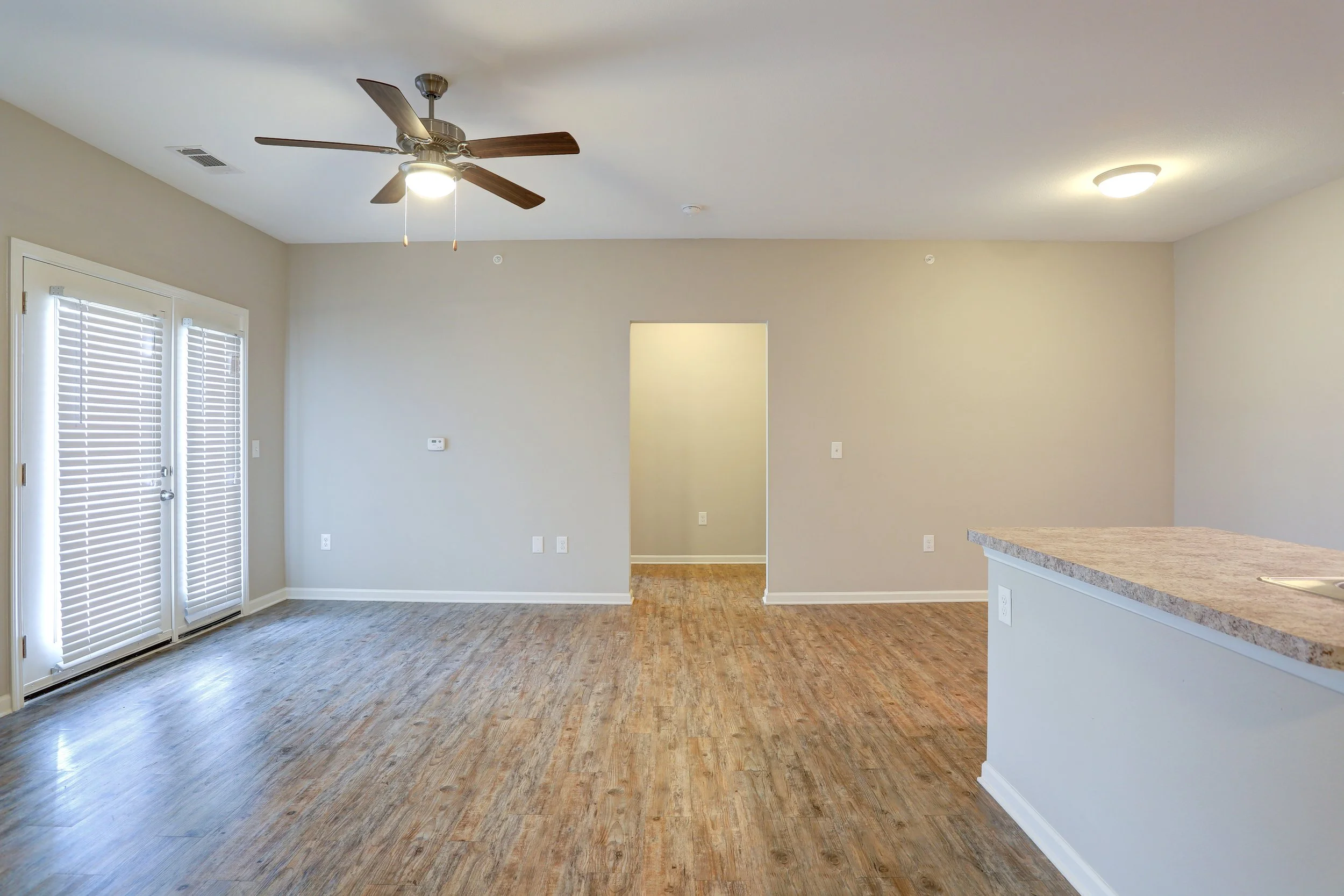 Interior of an empty living room with hardwood floors, a ceiling fan, a doorway, and a countertop in the kitchen area.