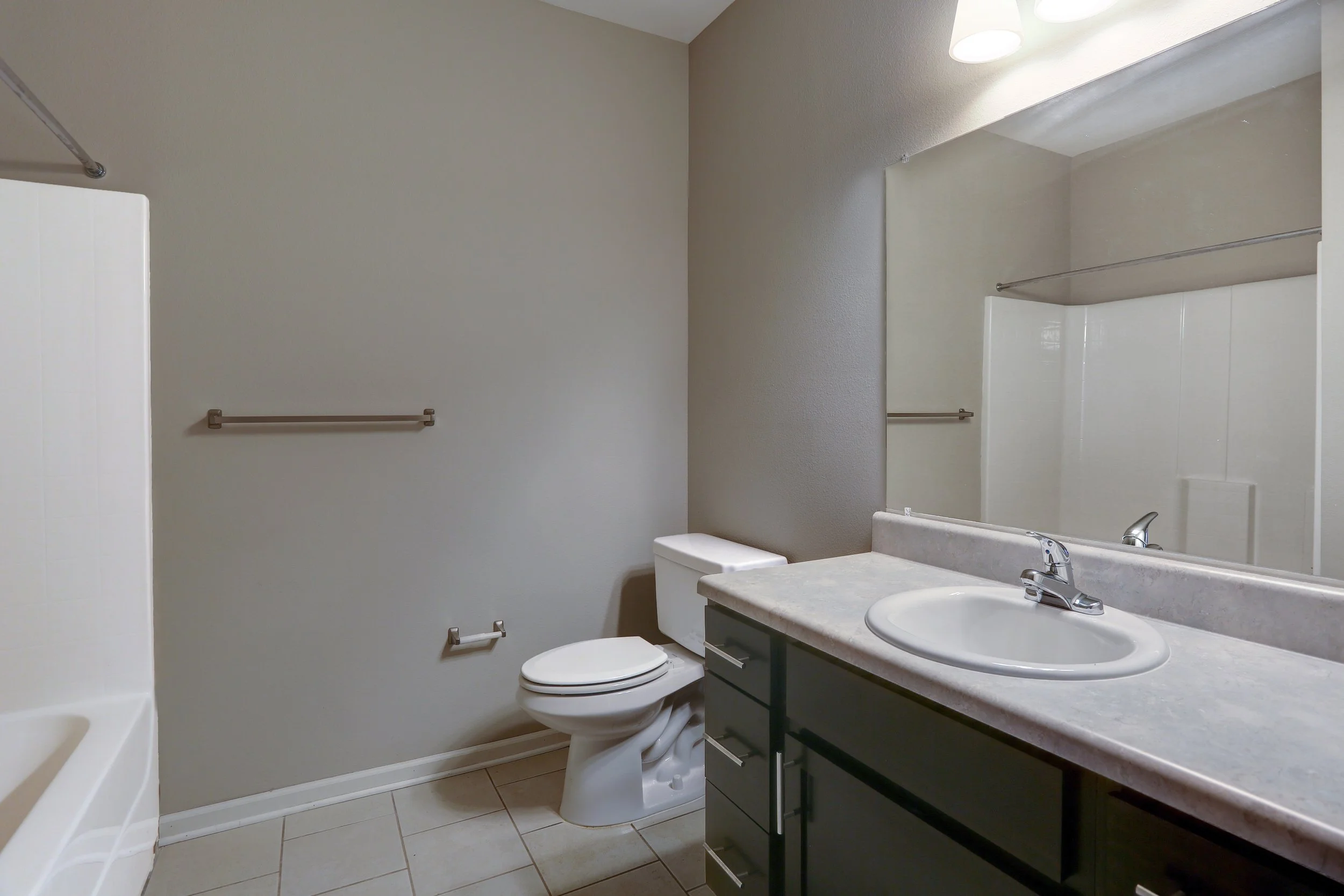 A small bathroom with a white bathtub, a toilet, a dark gray vanity with a marble countertop, a sink, and a large mirror. The walls are painted light gray, and there are beige tile floors. A metal towel bar is installed on the wall beside the bathtub