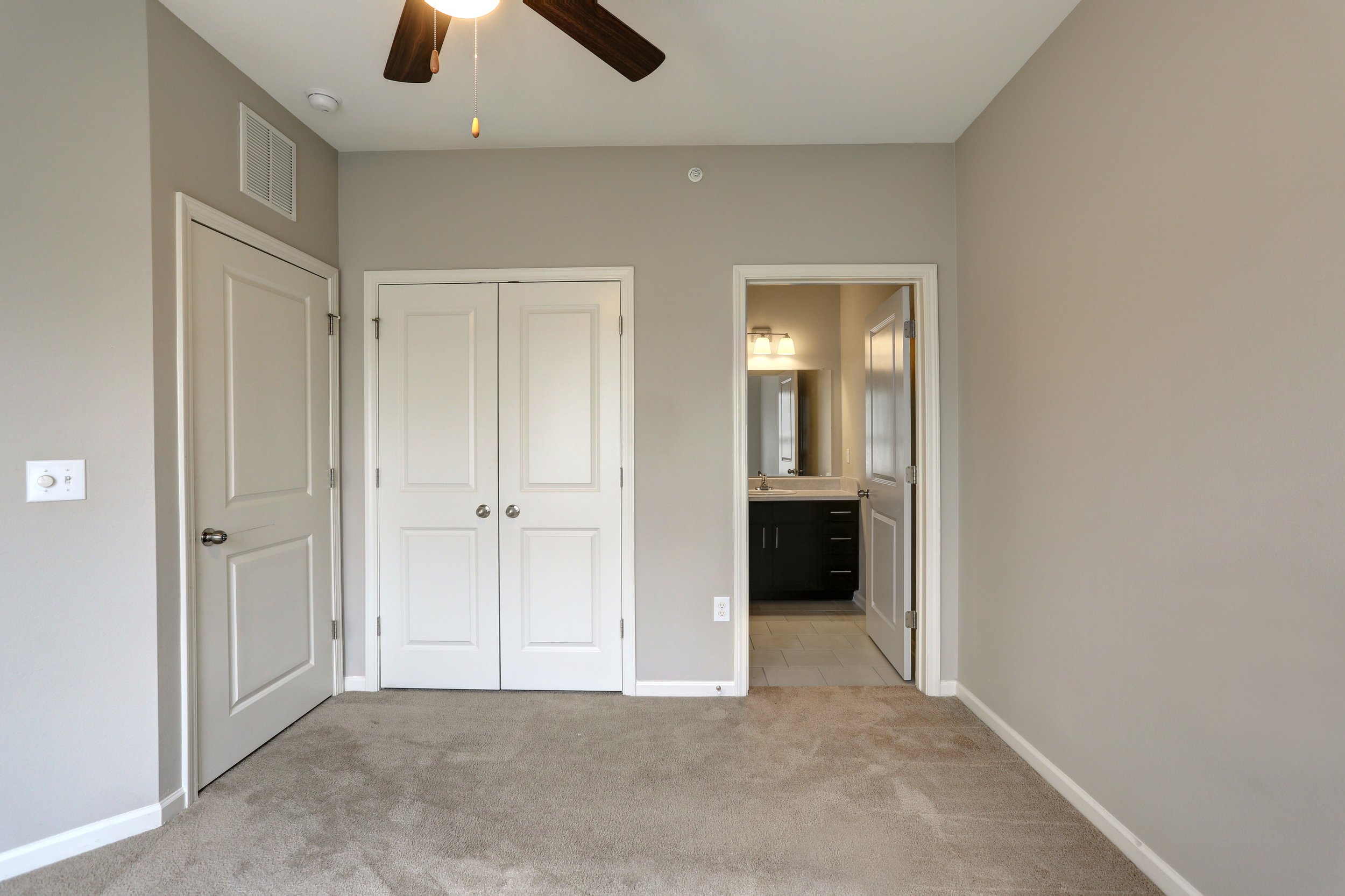 Empty bedroom with beige carpet, light gray walls, a ceiling fan, and a bathroom with a dark vanity and mirror.