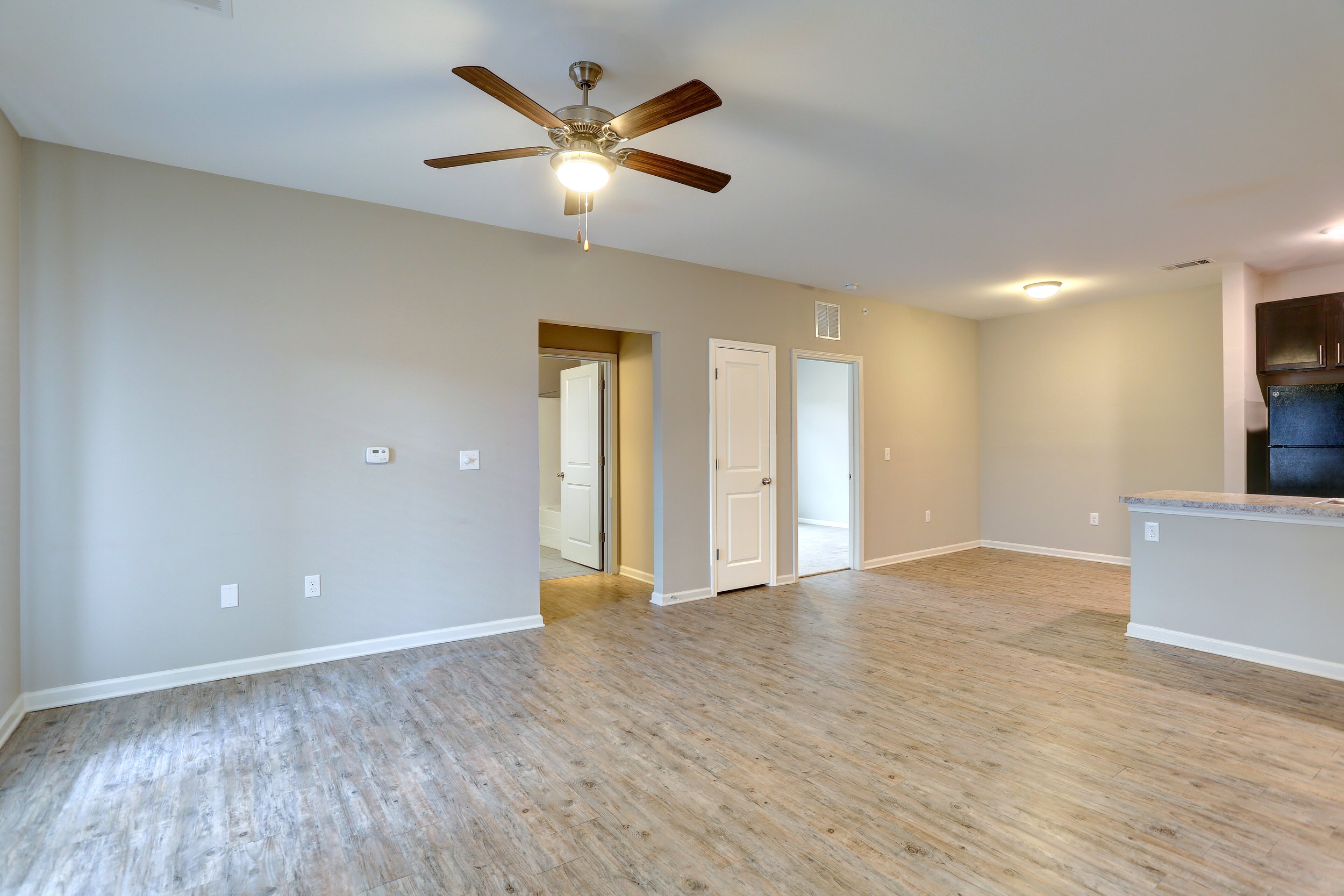 Empty living room with light beige walls, wooden flooring, ceiling fan with light, and open doorways leading to other rooms.