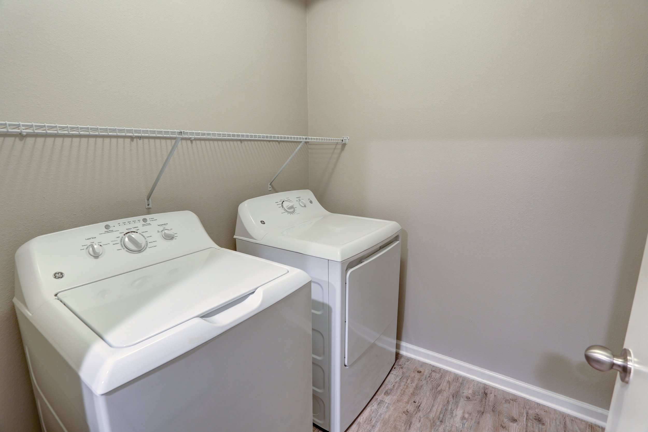 A laundry room with a white top-loading washing machine and a front-loading dryer, placed side by side against a beige wall with a wire shelf above them. The floor is wood-patterned vinyl, and a door with a round doorknob is partially visible on the 
