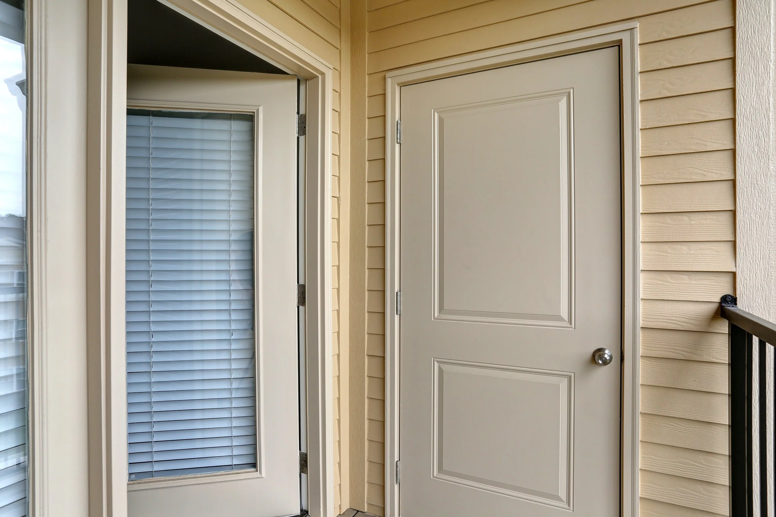 A front porch door with white trim, a peephole, and a round doorknob, next to a window with white horizontal blinds, on beige siding.