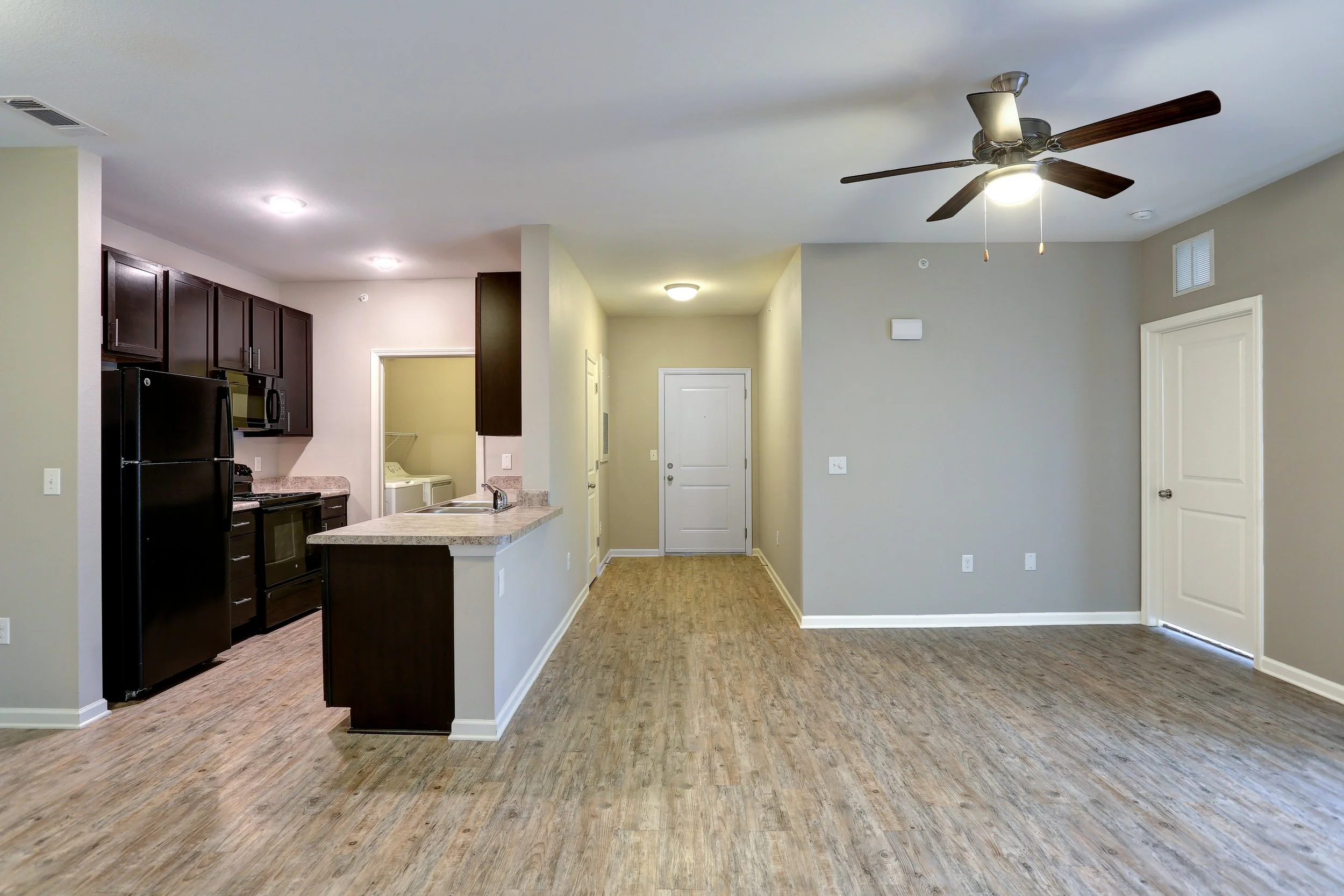 Interior view of a modern apartment with a kitchen area on the left, featuring dark cabinets, black appliances, and a granite countertop, and an open living space with wood flooring, light-colored walls, and a ceiling fan.