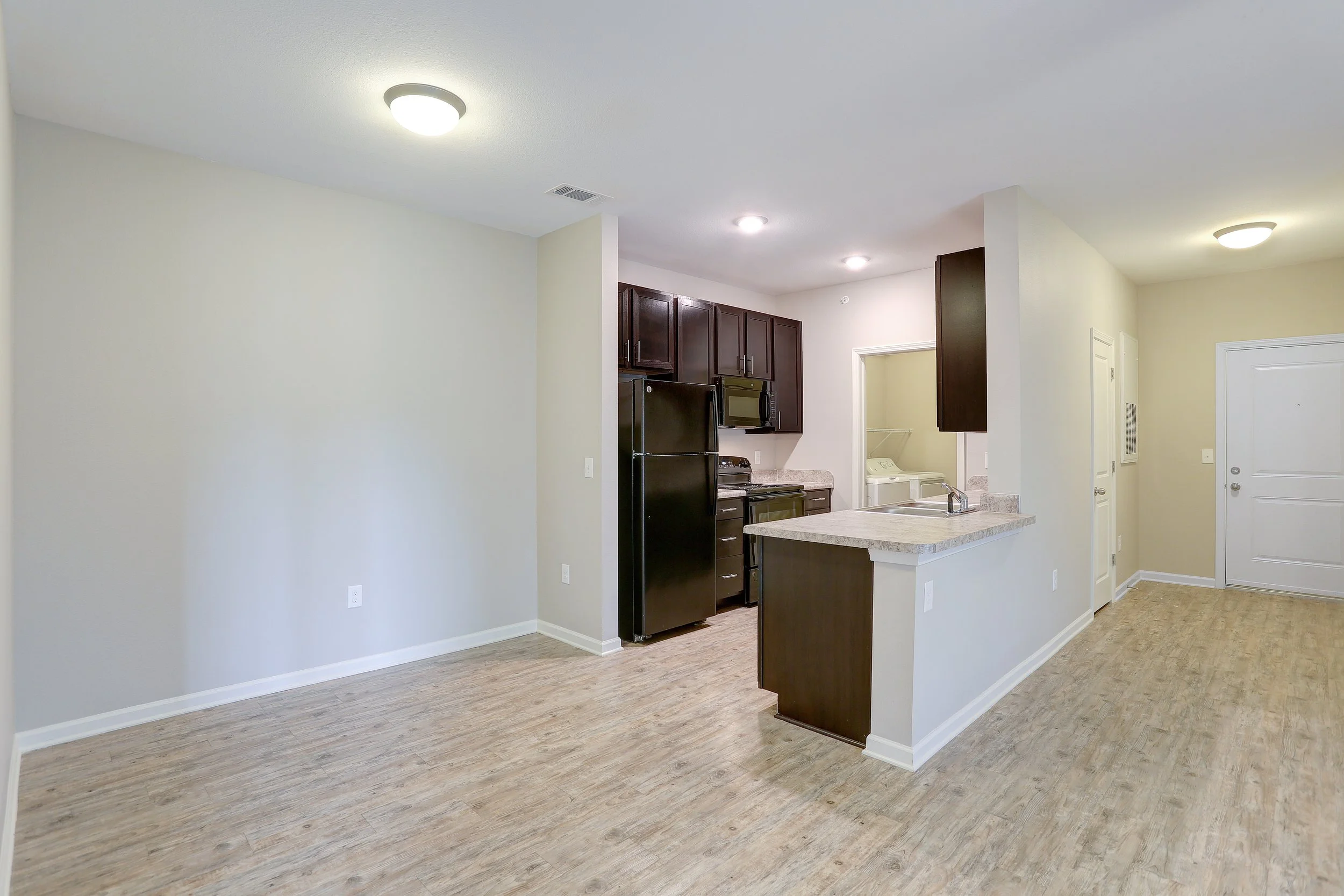 Interior view of a modern apartment with an open kitchen featuring dark wood cabinets, black appliances, and a granite countertop island. The living area has beige walls, wood flooring, and ceiling lights.