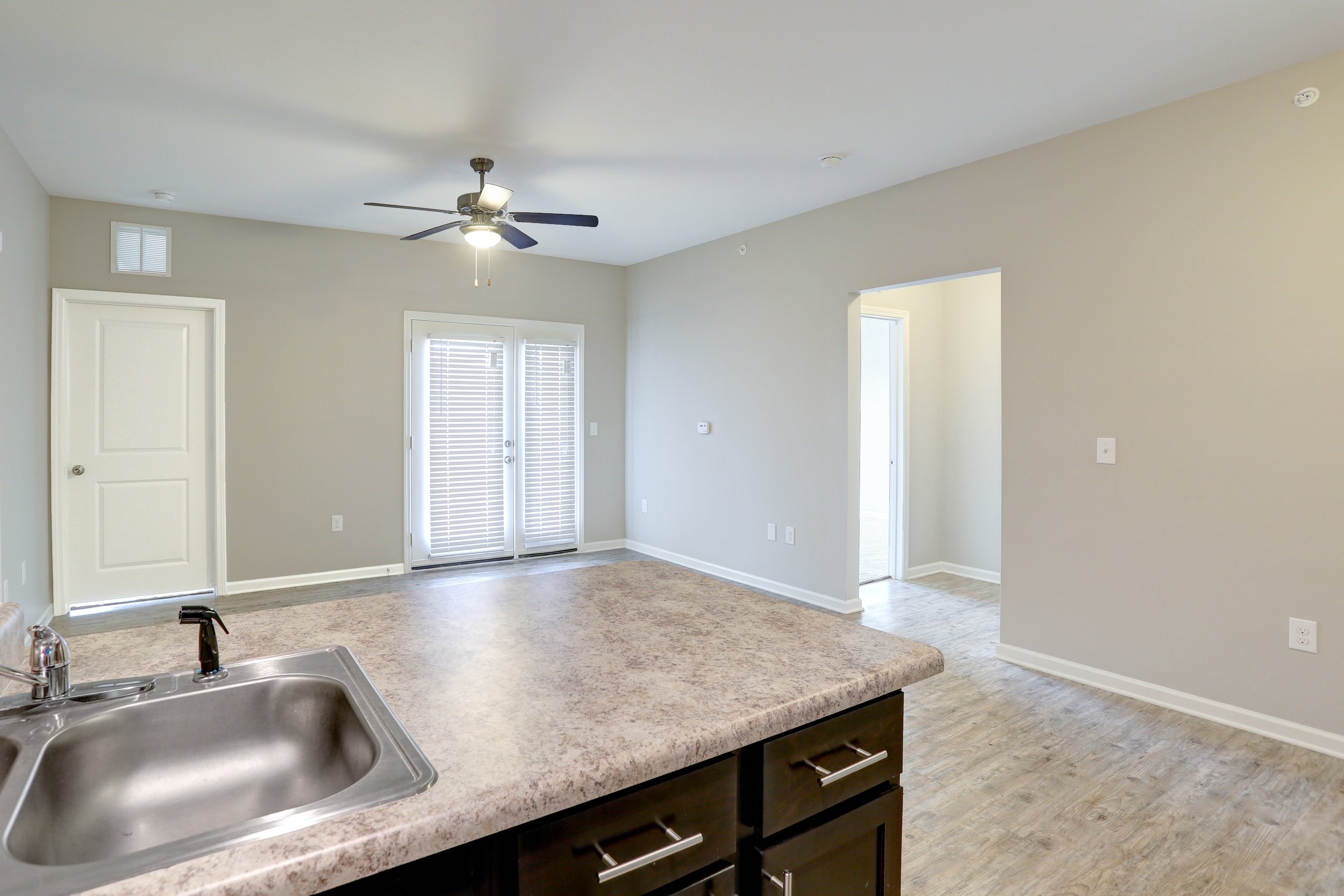 Interior view of a modern apartment with a kitchen island, stainless steel sink, light wood flooring, gray walls, white trim, a ceiling fan, and a door with blinds leading to a balcony or patio.