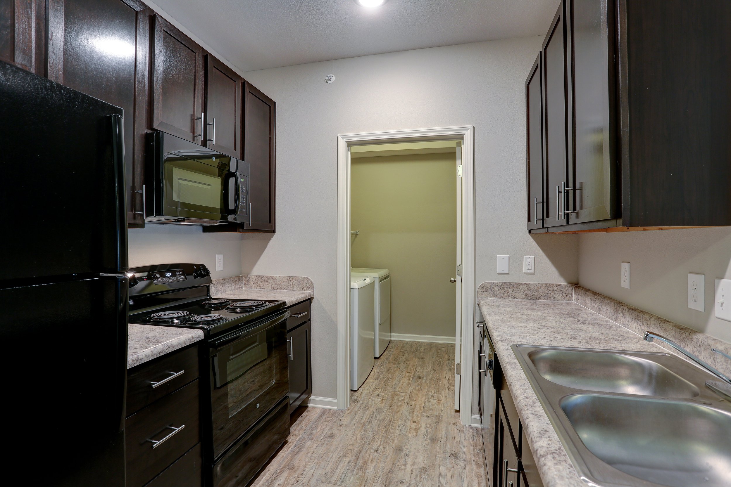 A compact kitchen with dark wood cabinets on the left and upper cabinets on the right. There is a black refrigerator on the left and a black stove below a microwave. The countertops are made of light-colored material, and a double sink is visible on 
