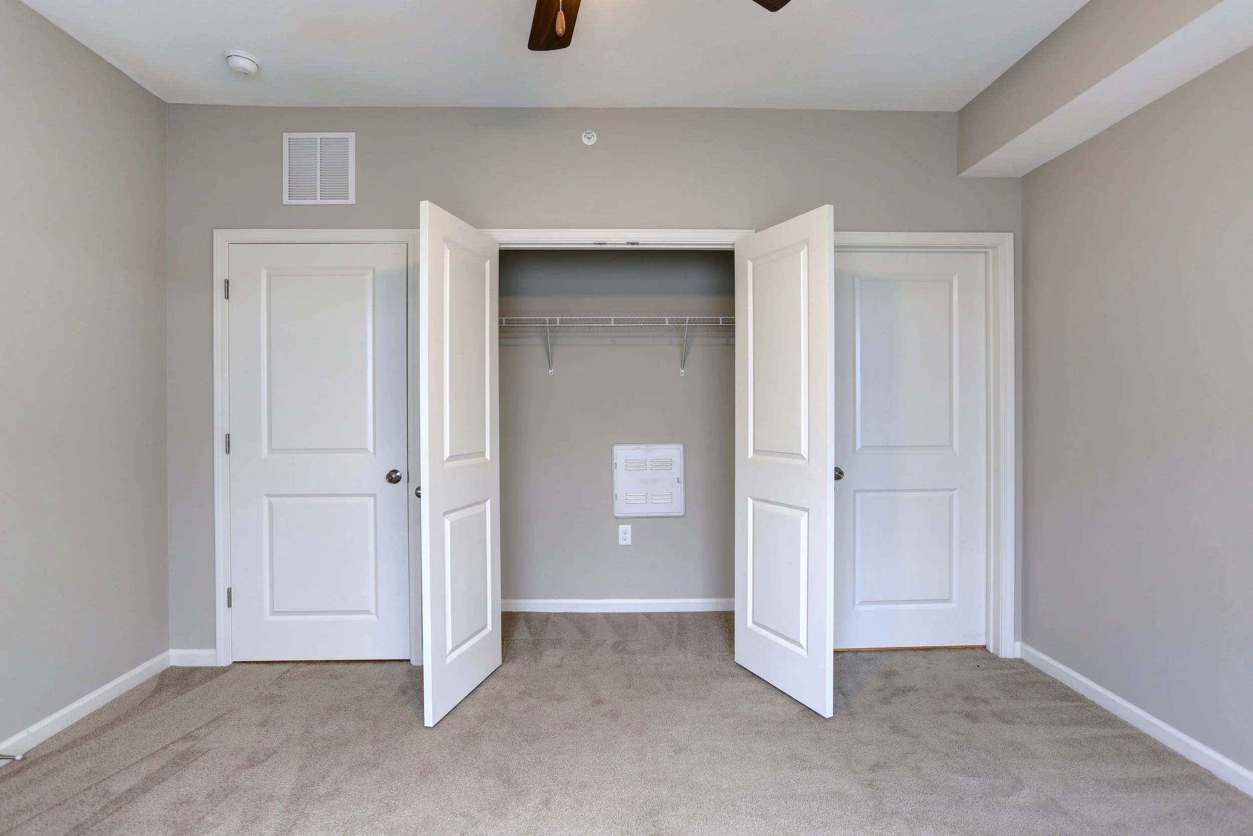 Empty closet with white bi-fold doors open, gray walls, beige carpet, and a ceiling fan visible at the top.