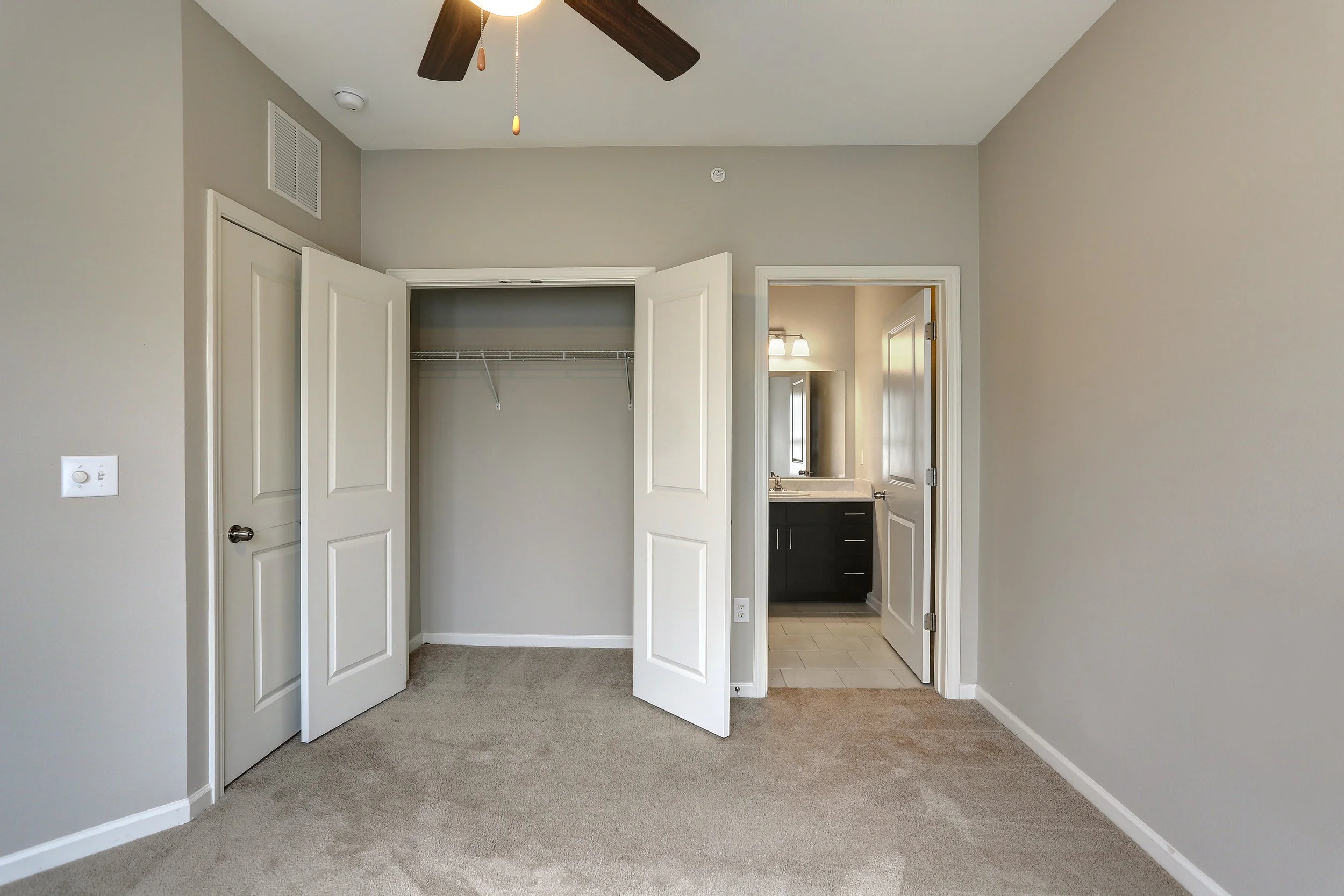A bedroom with open closet doors showing an empty closet with a wire shelf. There is a ceiling fan with dark blades and a light in the room. An open door leads to a bathroom with a dark vanity and a light fixture above the mirror.