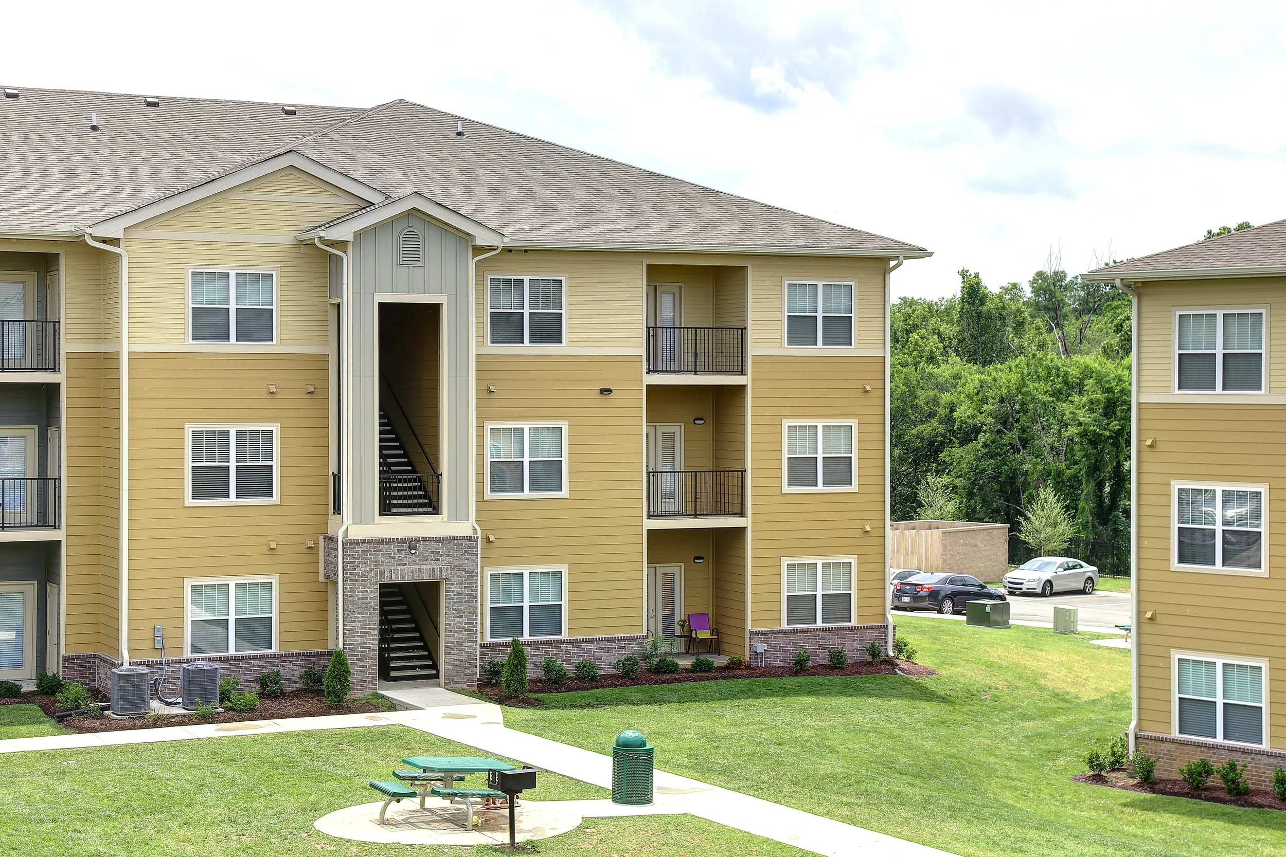 Apartment building with multiple floors, yellow siding, and balconies, surrounded by green lawn and trees, with parked cars in the background.