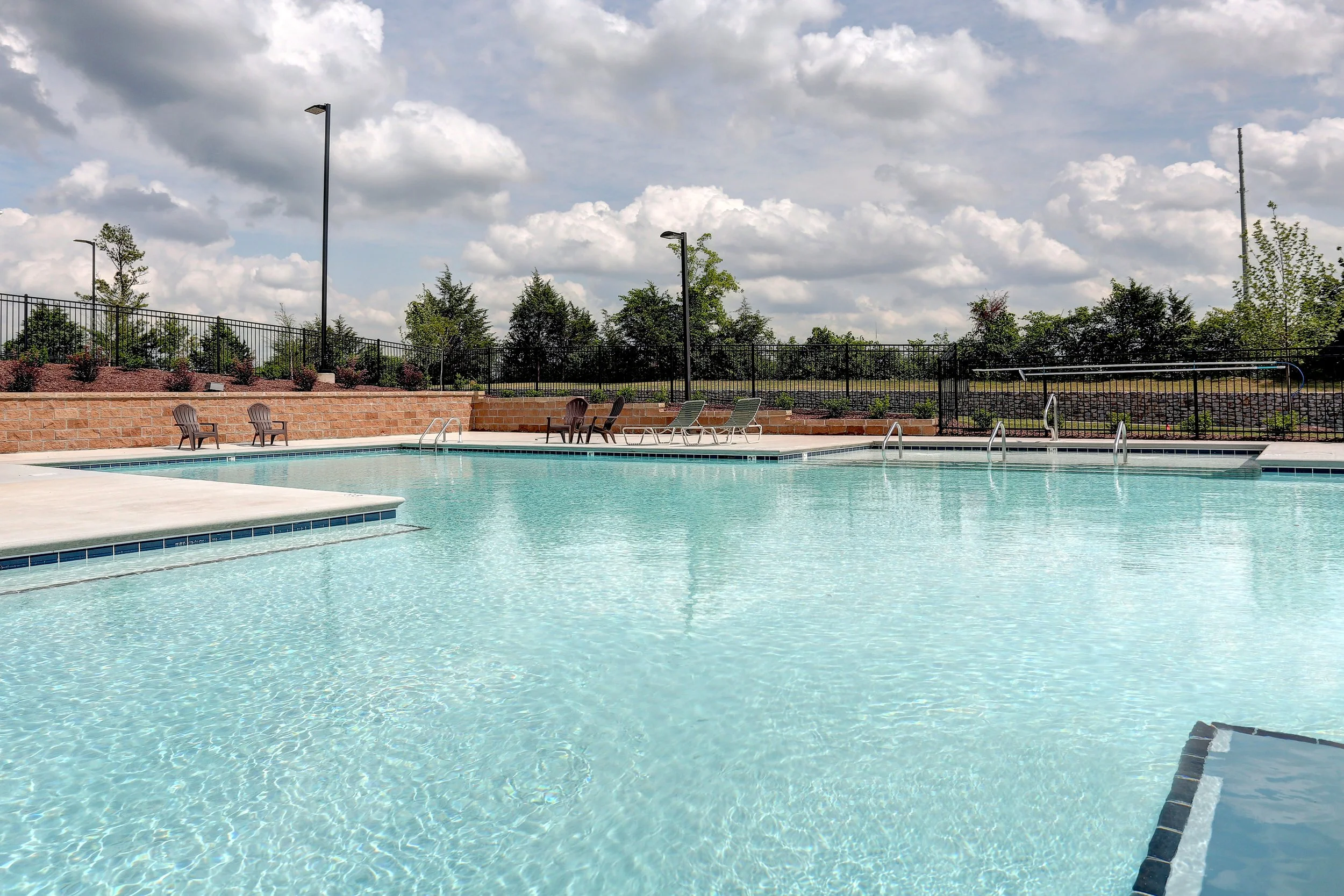 An outdoor swimming pool with chairs and a brick wall, surrounded by trees and a black fence under a cloudy sky.