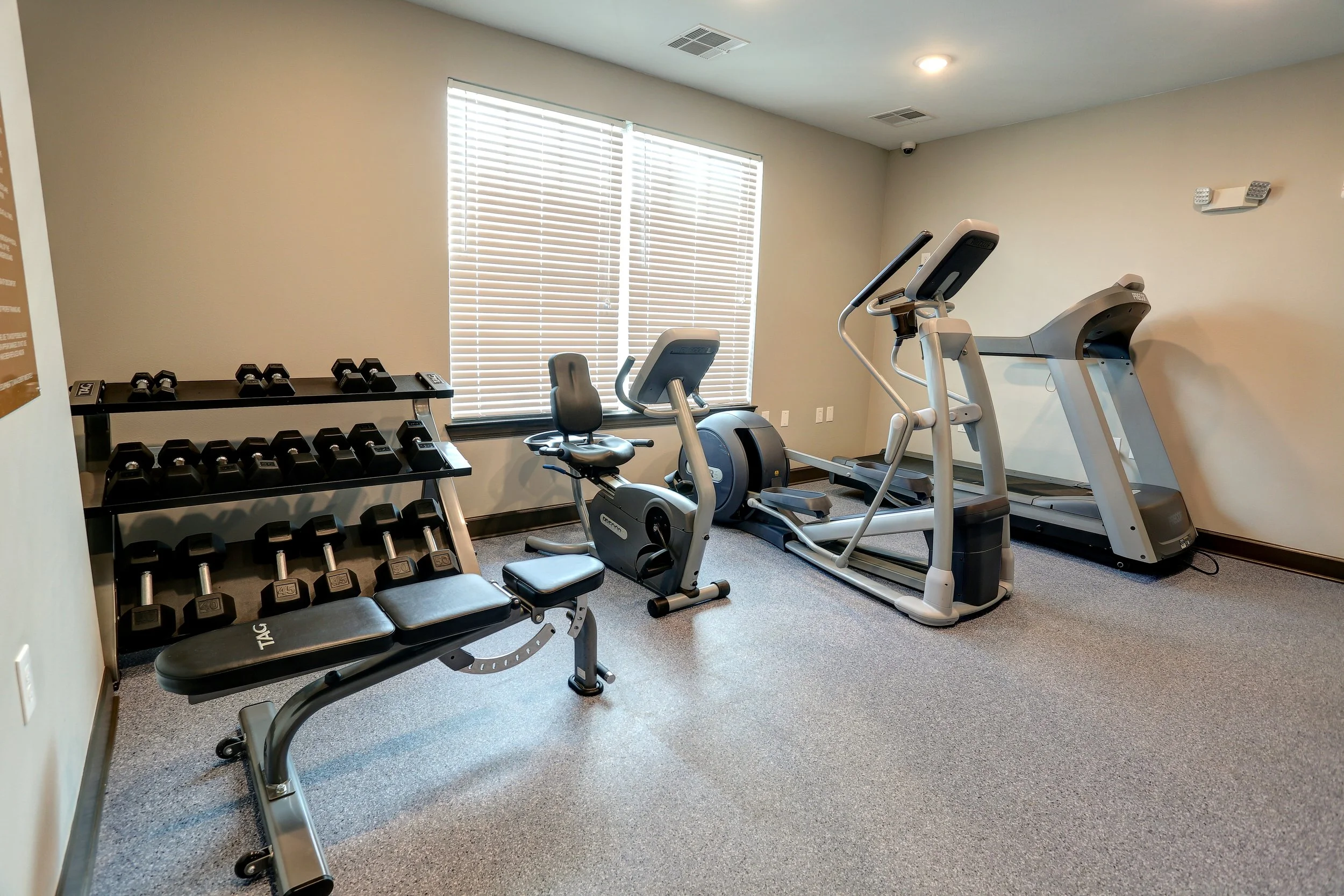 A small exercise room with a window with blinds, a stationary bike, a treadmill, a weight bench with dumbbells, and a small table for weights.