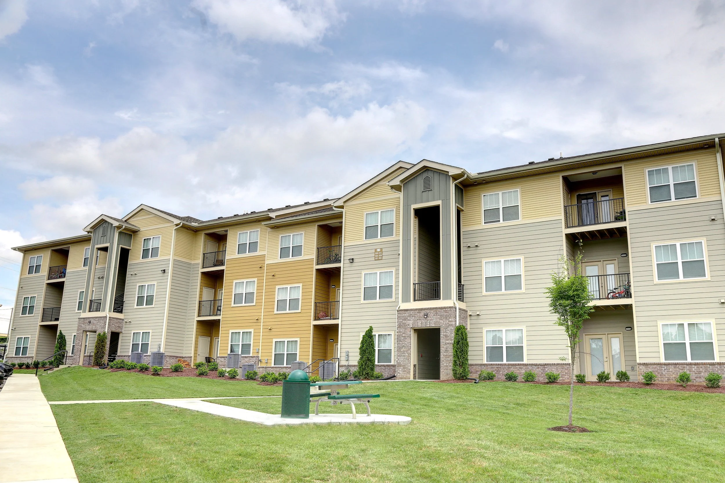 A multi-story apartment building with beige and gray siding, balconies, and a landscaped lawn with trees, a sidewalk, a bench, and a trash can in front.