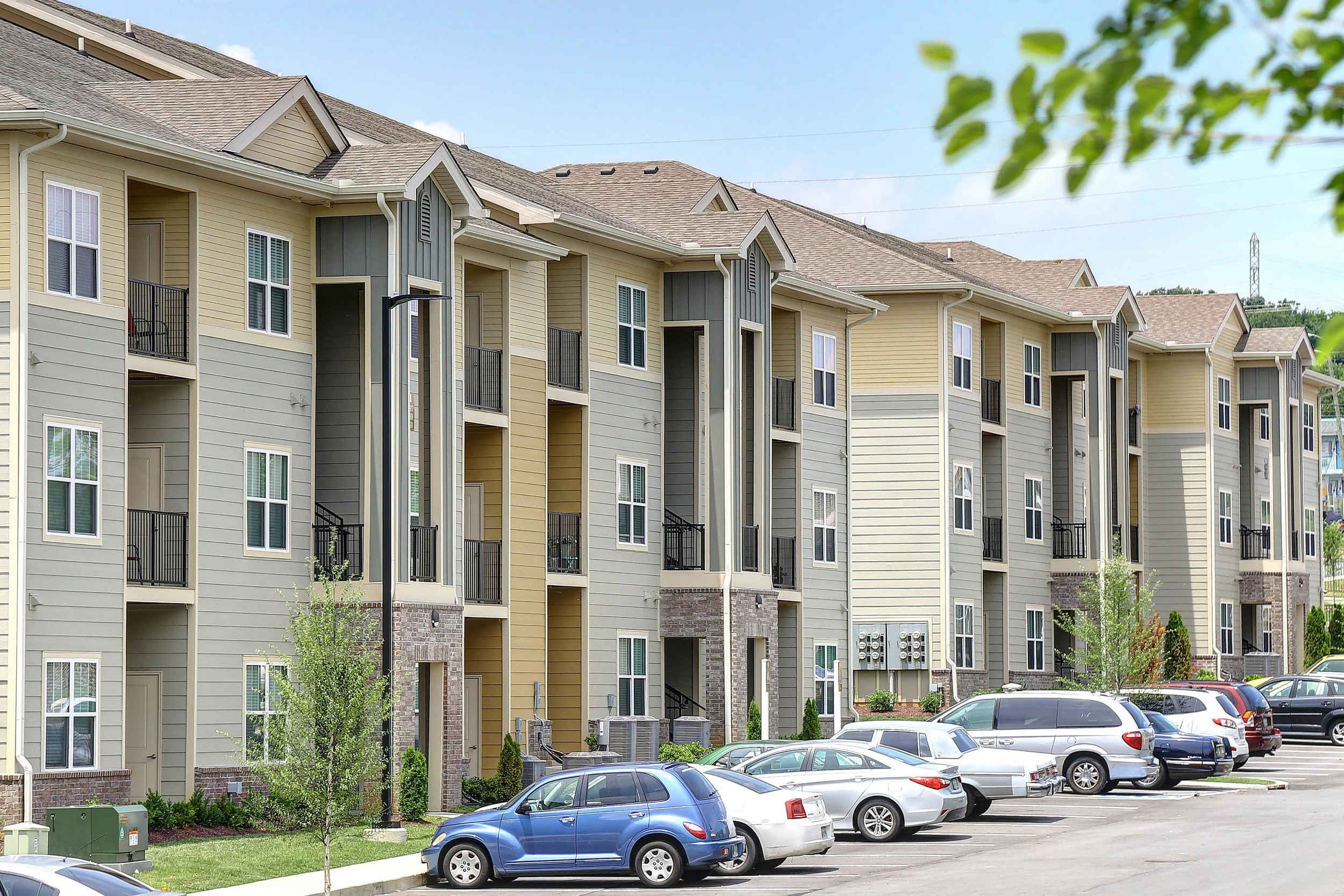 View of a multi-story apartment complex with beige and gray siding, multiple balconies, and windows, with a parking lot filled with cars in the foreground and green trees nearby.