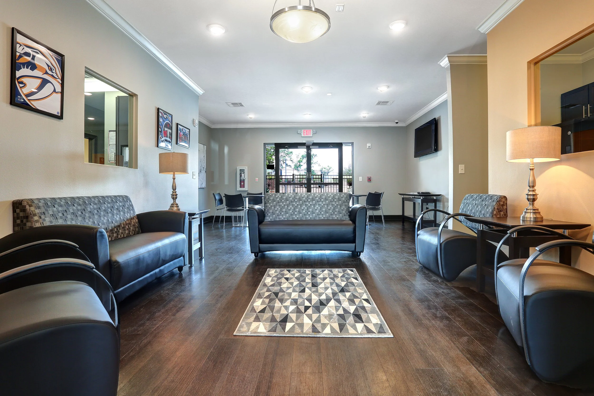 A spacious waiting room with black leather armchairs and a patterned rug on a dark wood floor. Light beige walls are decorated with framed artwork, and a large window with glass doors leads outside. There are lamps on side tables and a wall-mounted T