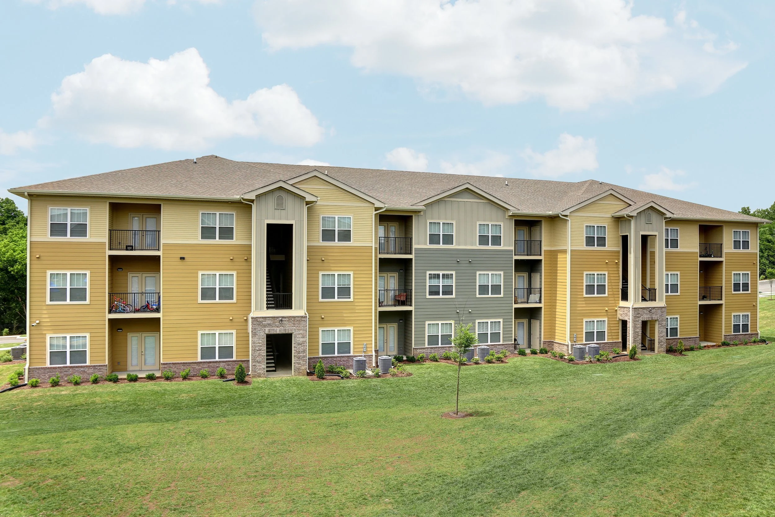 An apartment building with yellow siding, green siding, and stone accents, surrounded by a grassy lawn with a small tree and landscaping.