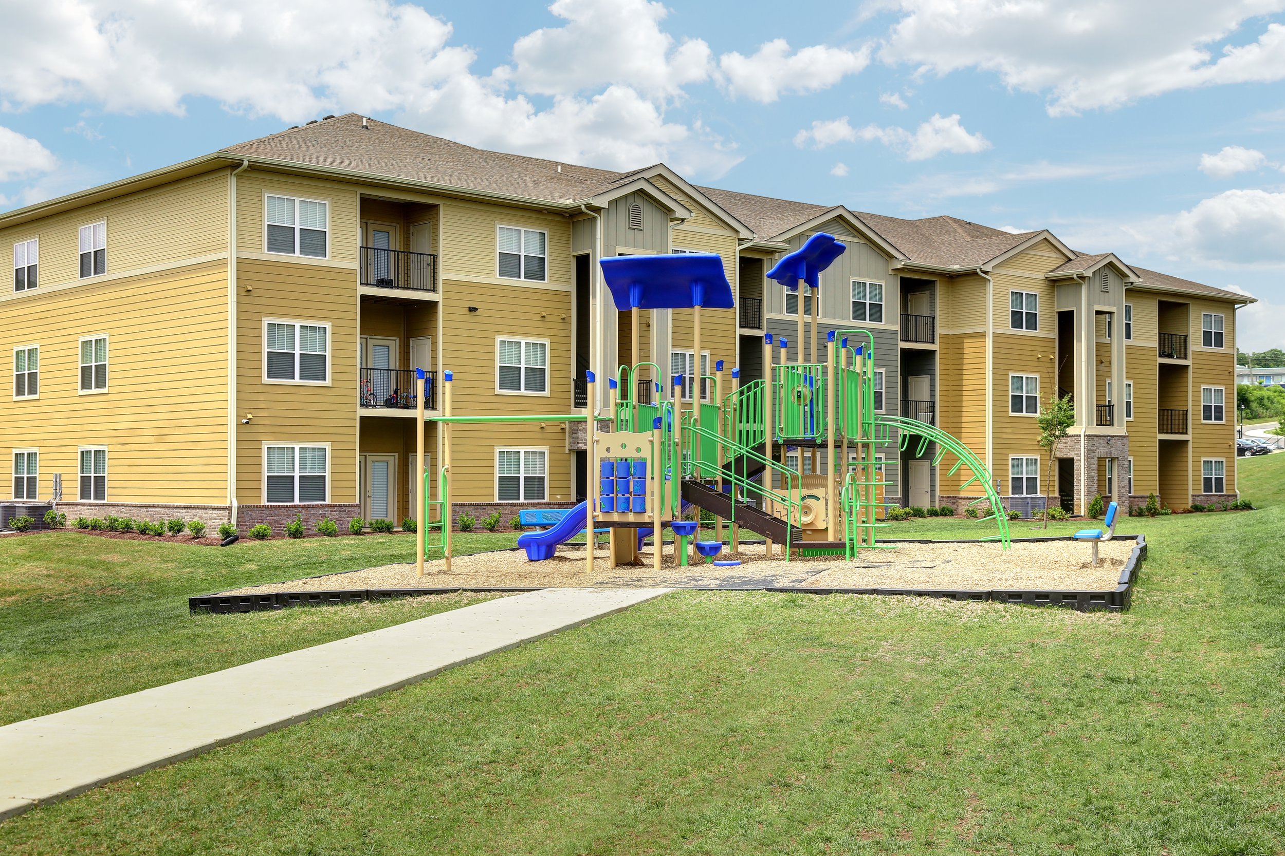 An apartment complex with yellow and gray siding, multiple balconies, and a playground with blue, green, and black equipment in the foreground. The grass is well-maintained, and a sidewalk runs through the area, under a partly cloudy sky.