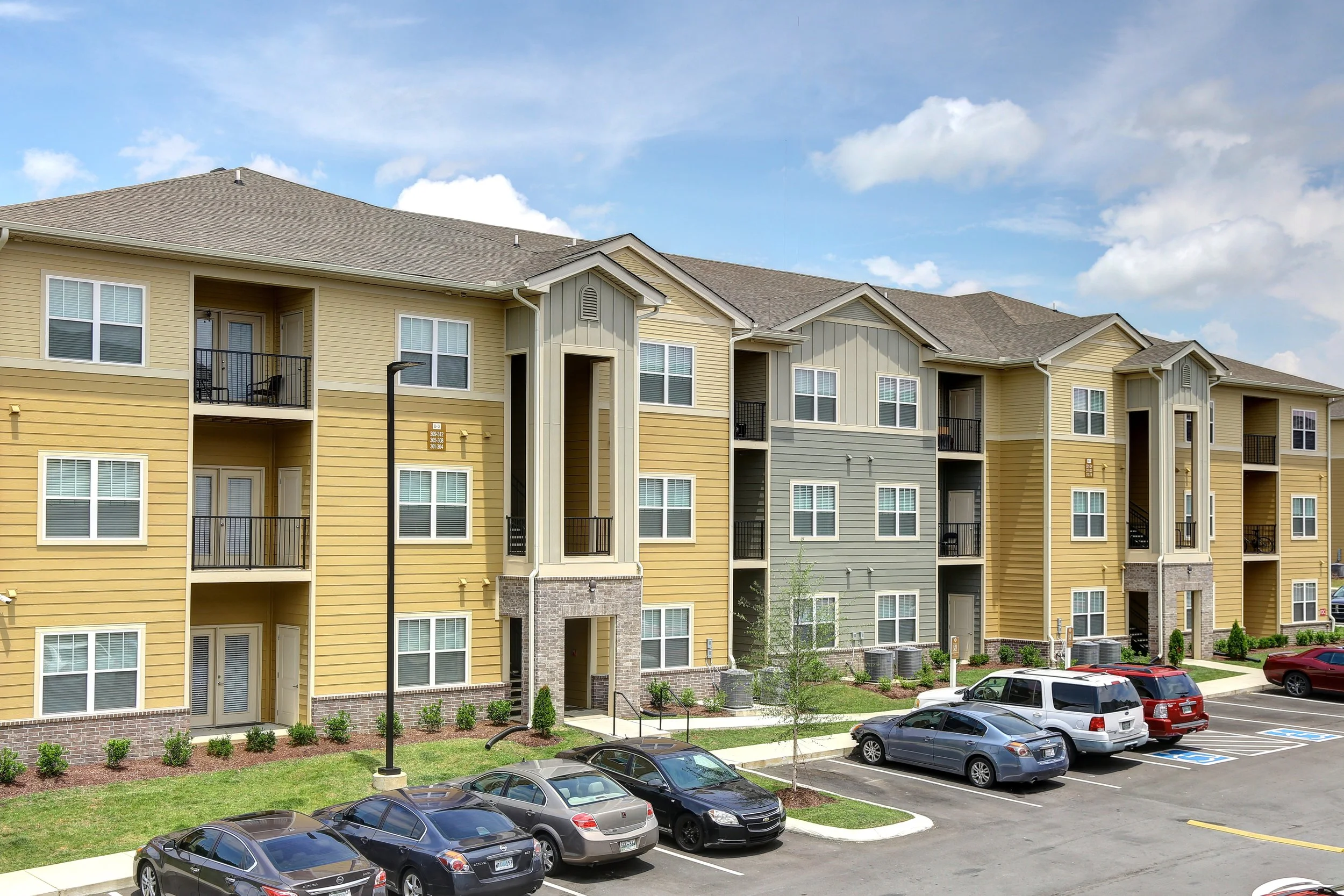 A multi-story apartment complex with yellow and gray siding, multiple balconies, and an adjacent parking lot with several parked cars under a partly cloudy sky.
